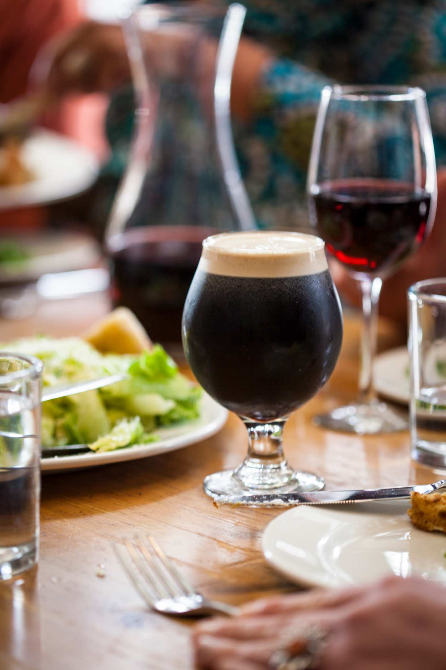 A glass of dark beer, a wine glass, and a plate of salad are arranged on a wooden table, with a blurred background suggesting a cozy dining setting.