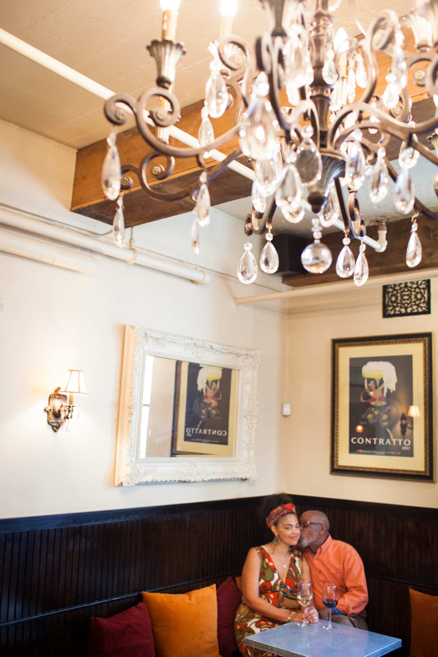 The image shows a cozy and intimate setting, with a couple seated at a table in the foreground, surrounded by a decorative chandelier and framed artwork on the walls in the background.
