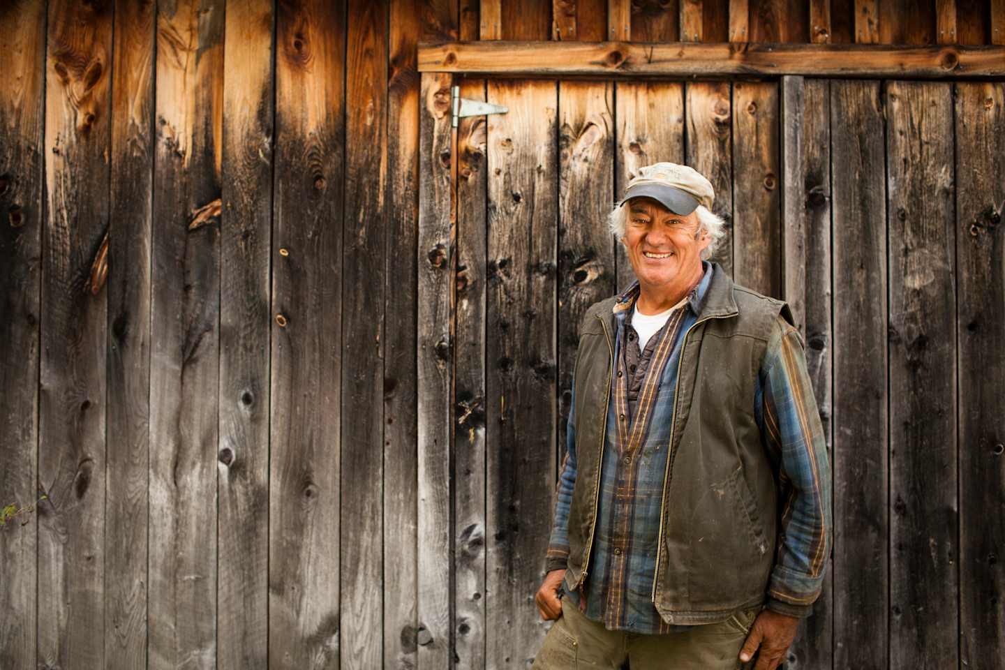 An elderly man wearing a cap and casual clothing stands in front of a weathered wooden wall, his expression conveying a sense of contemplation.