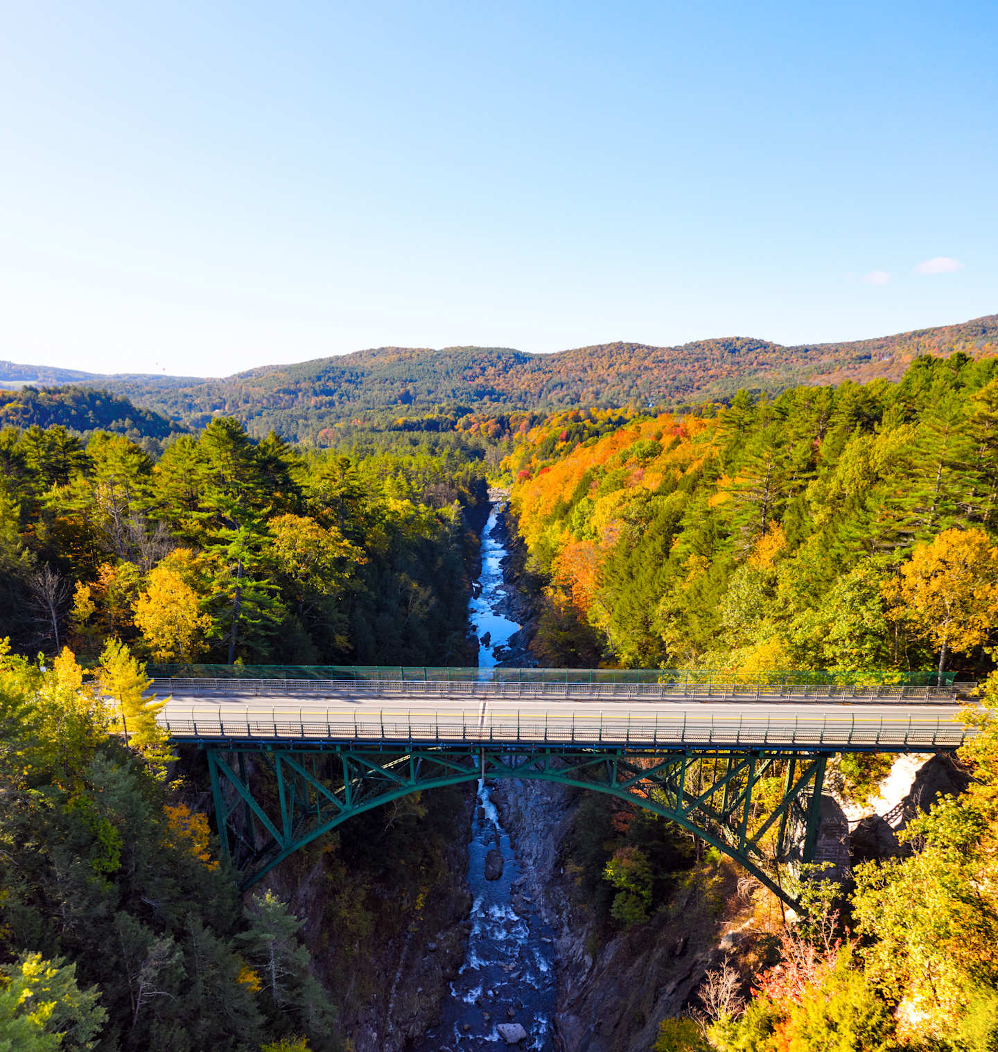 A scenic autumn landscape with a bridge spanning a deep gorge, surrounded by vibrant foliage-covered hills and mountains in the background.