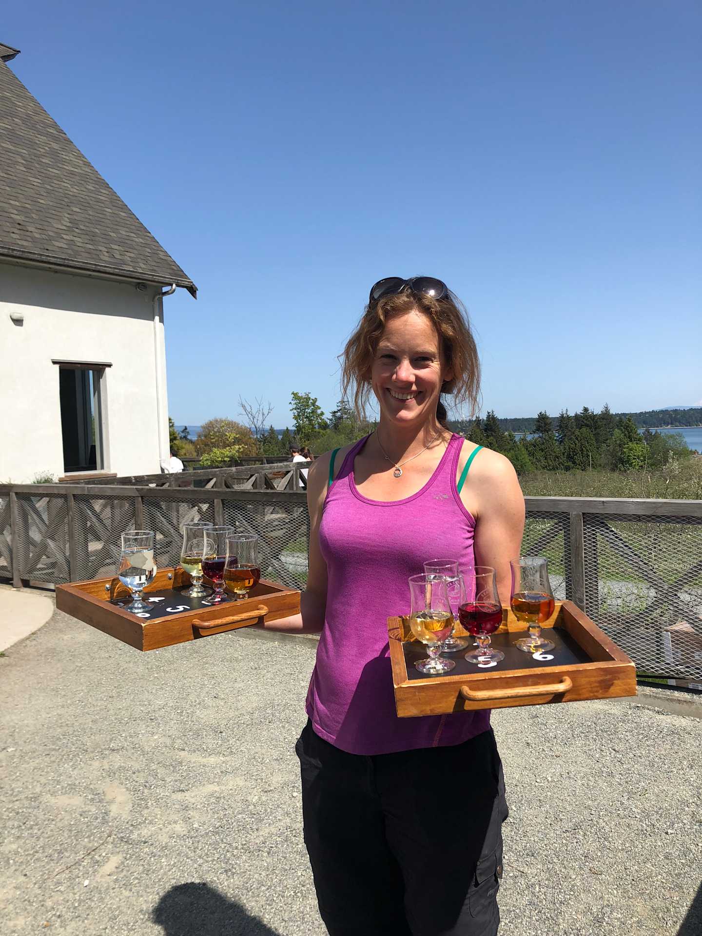 A young woman in a pink tank top is holding a tray of beer samples outdoors, with a wooden building and scenic landscape visible in the background.
