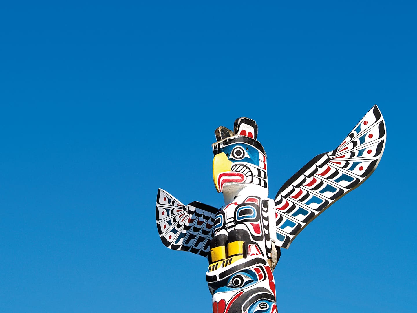 A colorful and intricately carved totem pole with a bird-like figure prominently displayed against a clear blue sky.