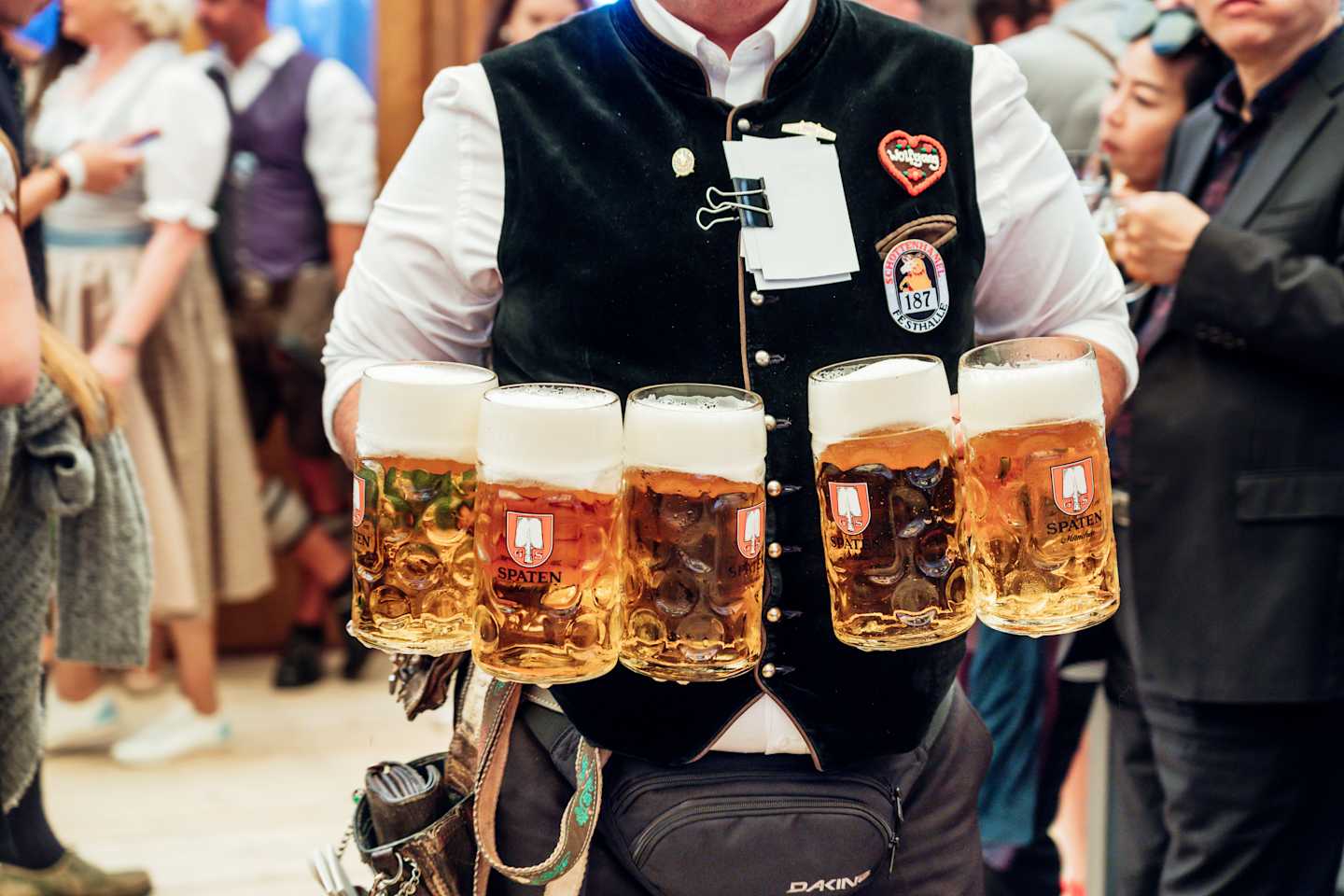 A person wearing a traditional Bavarian outfit is holding several large glasses of beer in a crowded indoor setting.