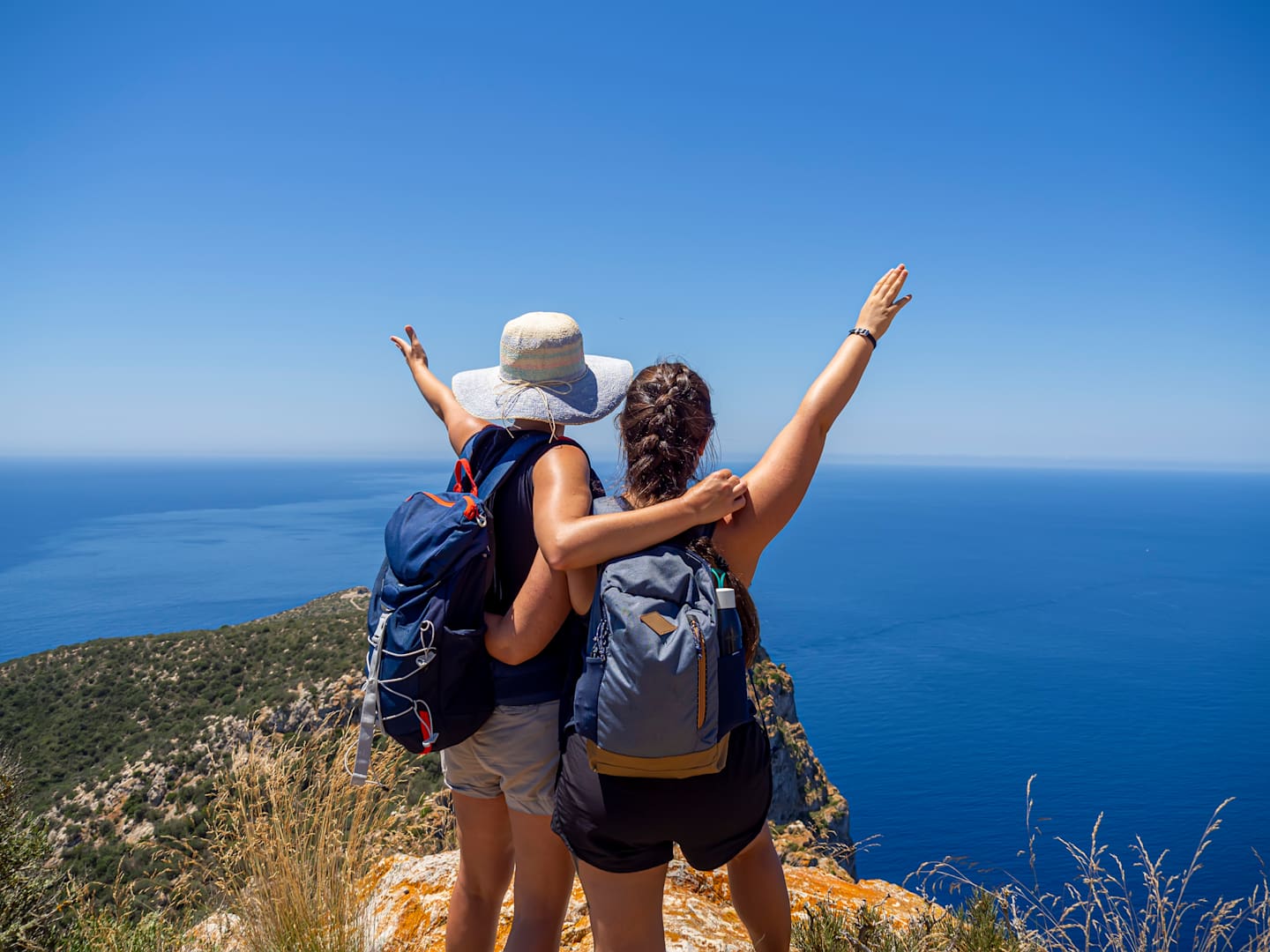 Two hikers with backpacks stand on a cliff overlooking a vast, blue ocean against a clear sky.