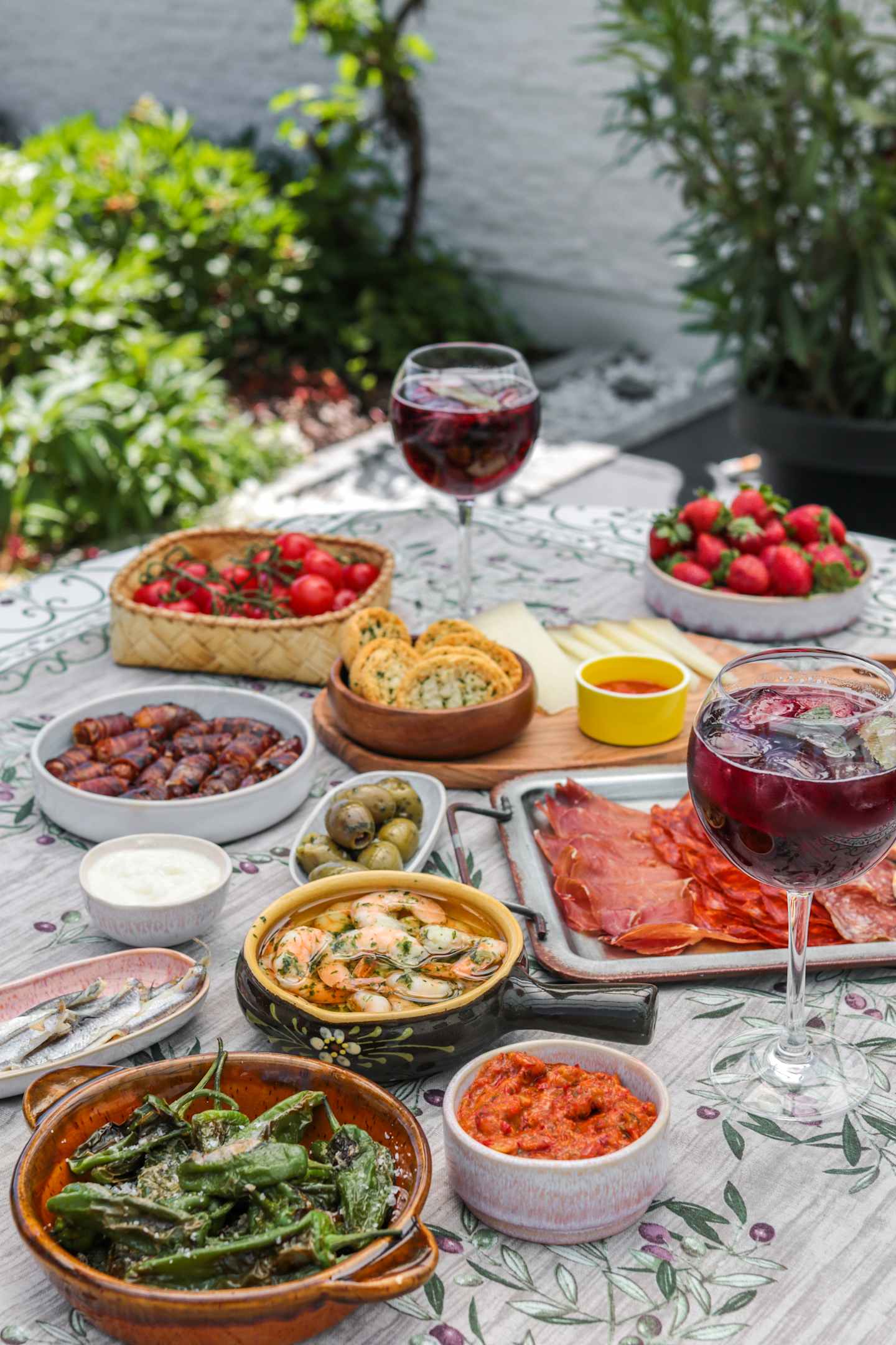 A table filled with various Mediterranean-style dishes, including a glass of red wine, fresh strawberries, and a variety of appetizers and main courses, set against a backdrop of lush greenery.