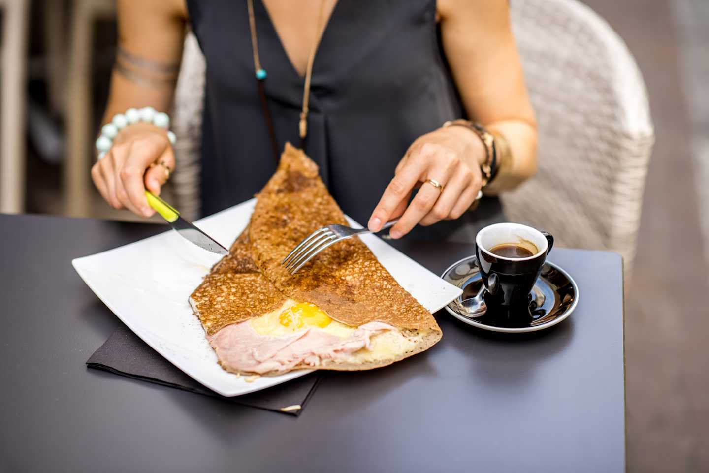 A person in a black top is sitting at a table, eating a pastry and drinking a cup of coffee.