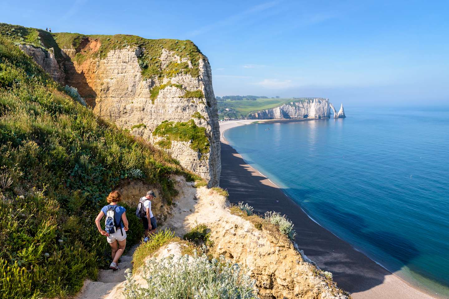 Rugged cliffs overlooking a tranquil blue sea, with two people hiking along a winding path in the foreground.