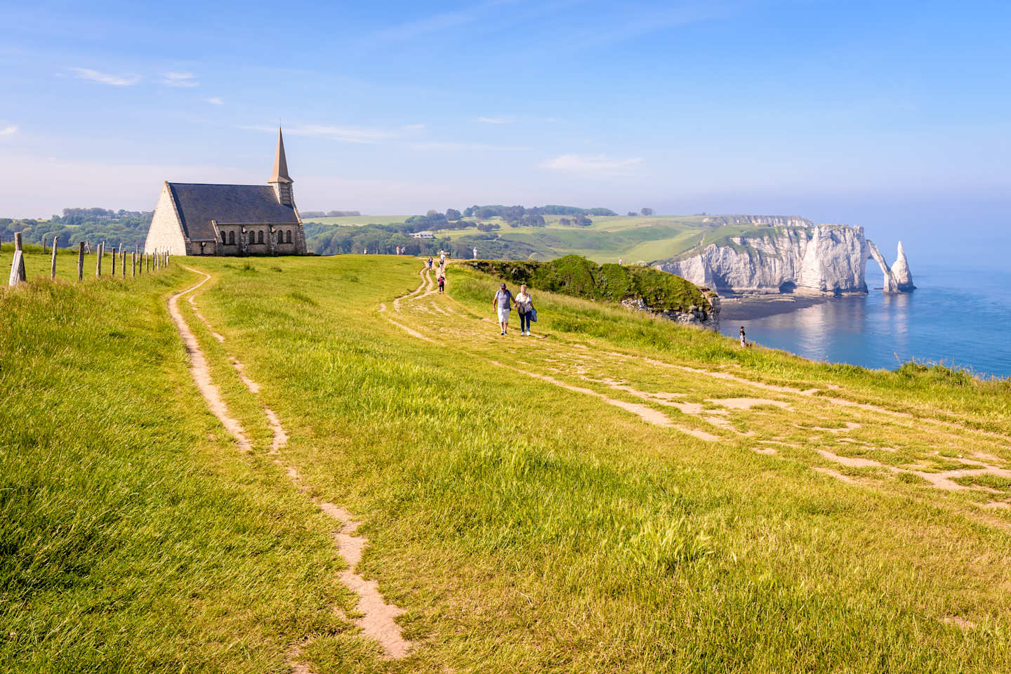 A winding path leads through a lush, grassy field towards a picturesque church, with the dramatic cliffs of the Normandy coast visible in the distance.