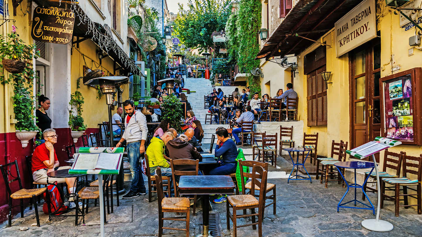 A vibrant and lively outdoor cafe scene with people dining at tables, surrounded by colorful buildings, greenery, and a bustling street in the background.