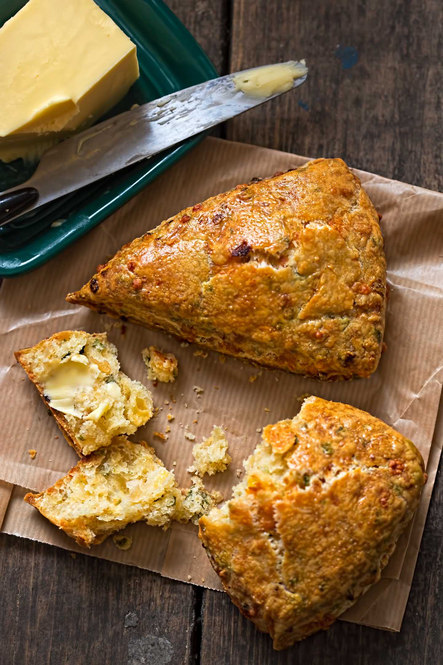 A golden-brown, freshly baked scone sits on a wooden surface, accompanied by a slice of butter on a green plate in the background.