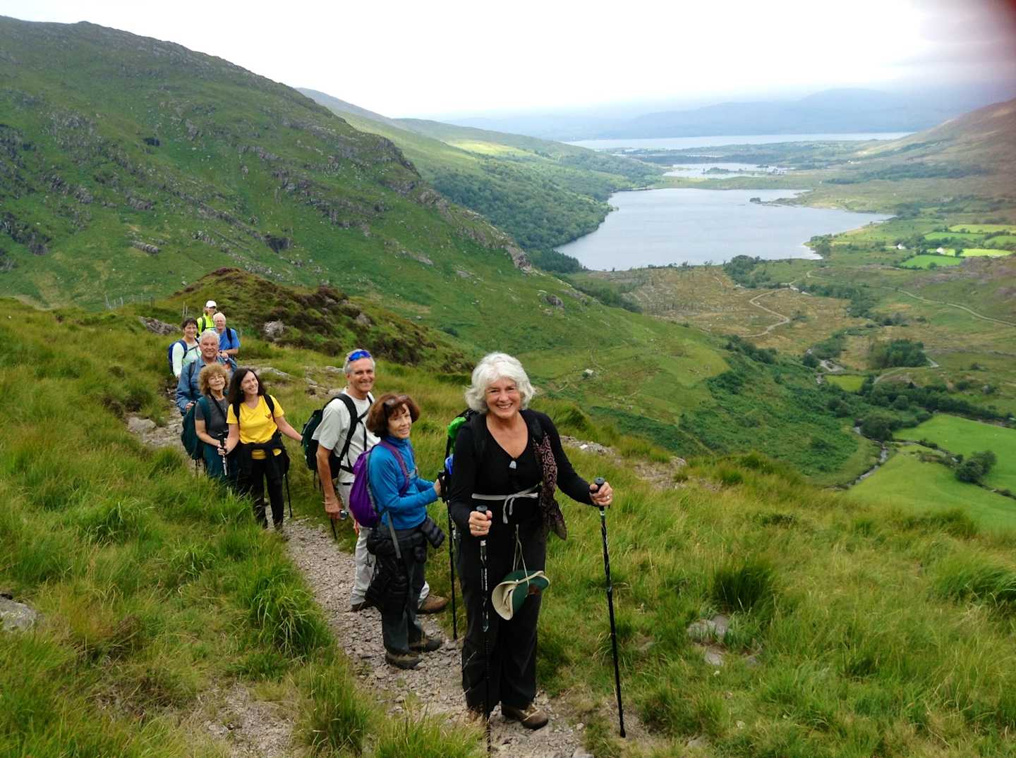 A group of hikers traversing a scenic mountain trail, with a picturesque lake and lush green hills visible in the background.