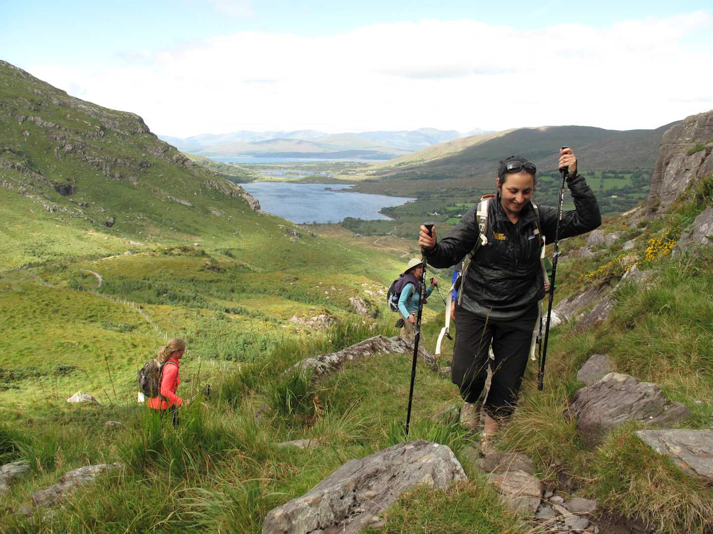 Two hikers are trekking on a trail through a lush, green landscape with mountains and a lake in the background.