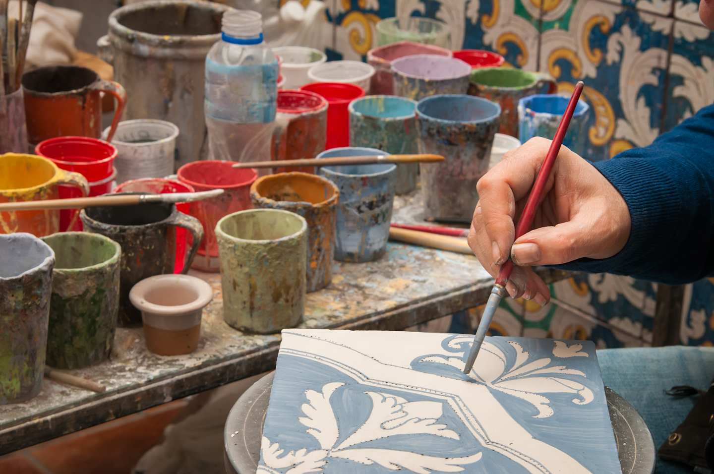 A hand holding a paintbrush is working on a decorative ceramic tile, surrounded by an assortment of colorful ceramic vessels and tools on a cluttered workbench.