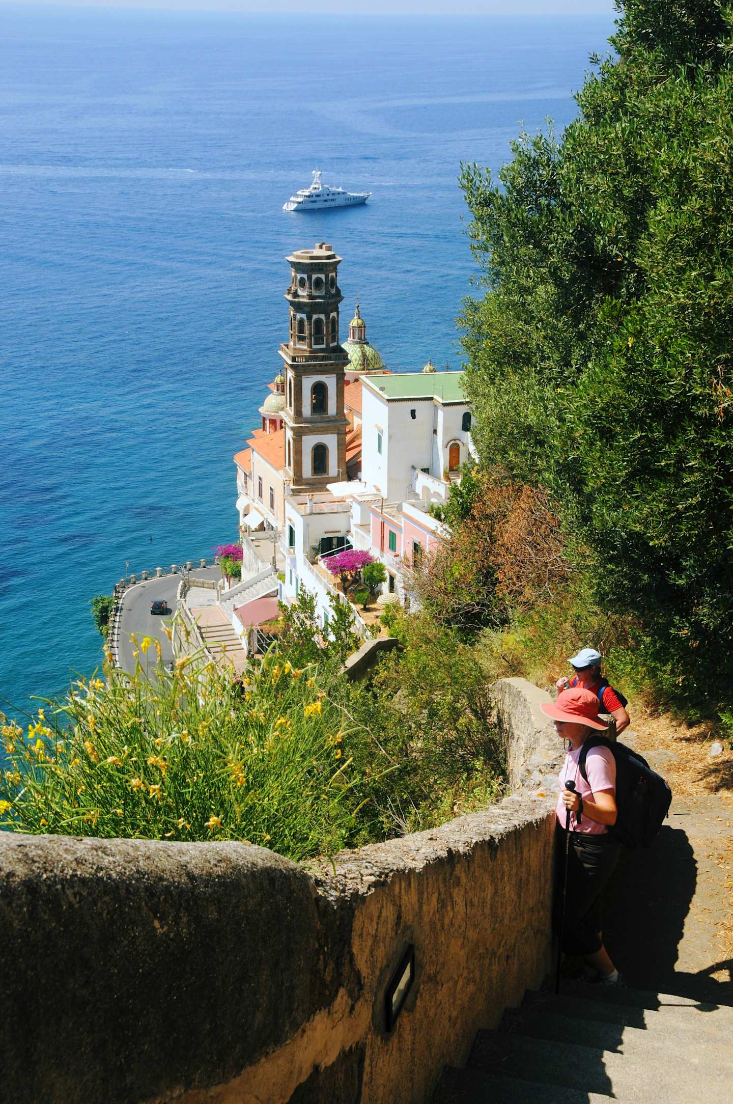 A picturesque coastal town nestled among lush greenery, with a colorful building and a cruise ship visible in the distance, while a person is seen walking along a winding path in the foreground.