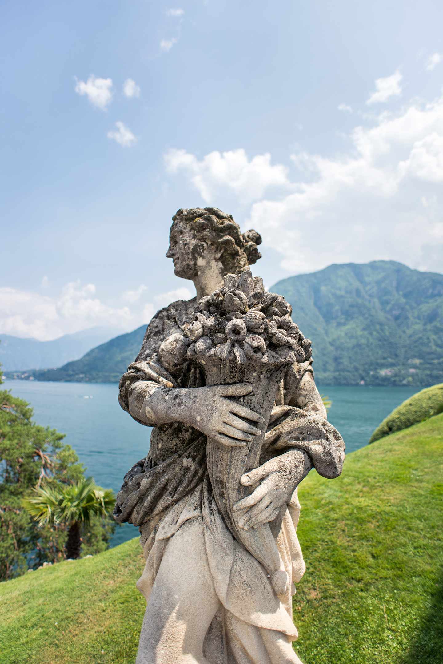 A stone sculpture of a figure embracing a large rock formation stands on a grassy hill overlooking a scenic lake and mountains in the background.