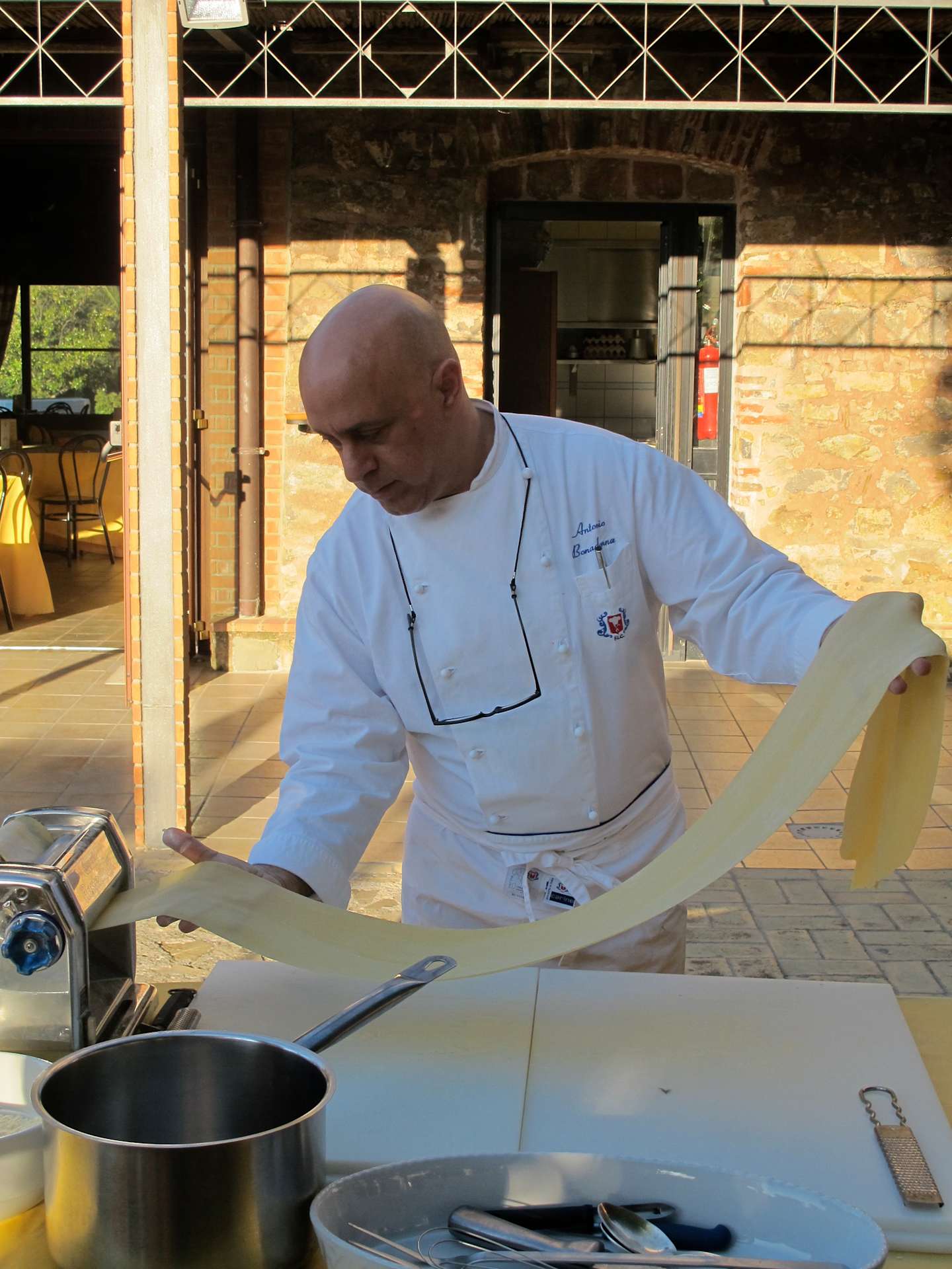 A chef in a white uniform is preparing food on a counter in an outdoor setting with a brick building in the background.