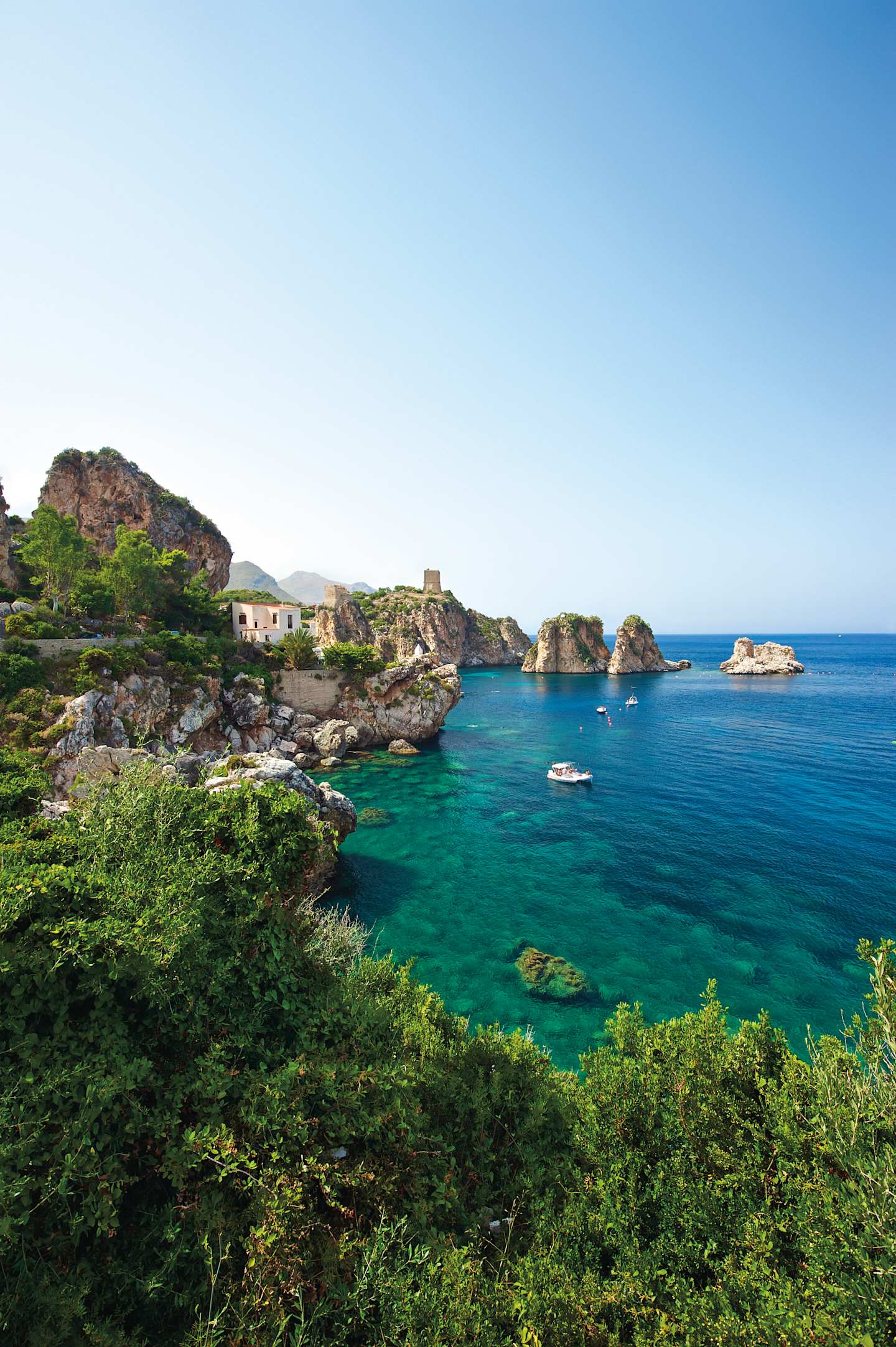 A picturesque coastal landscape with lush green vegetation in the foreground, rocky cliffs, and a serene turquoise sea in the background, under a clear blue sky.