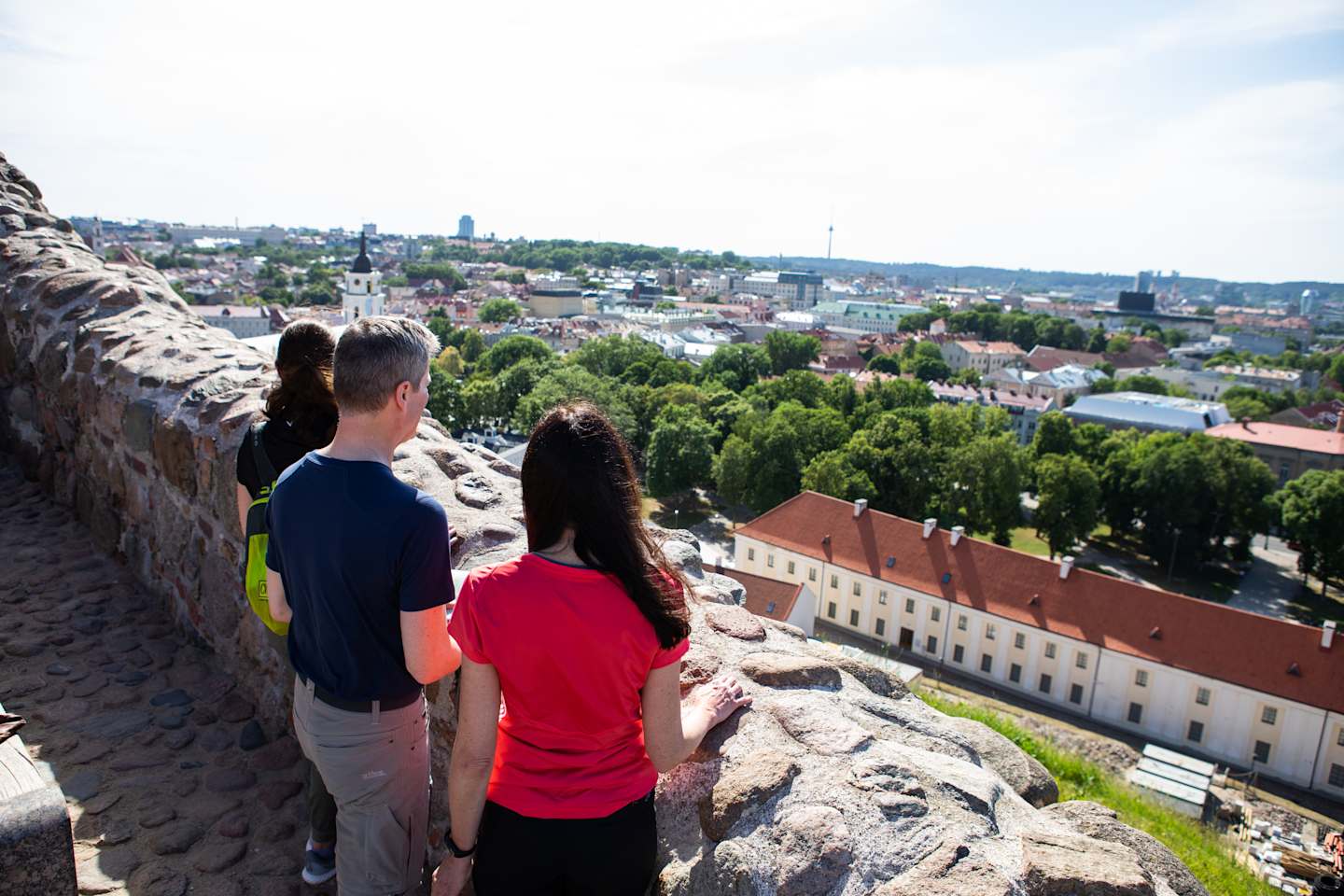 The image shows a couple standing on a rocky outcrop, overlooking a picturesque city with colorful buildings, churches, and greenery in the background.