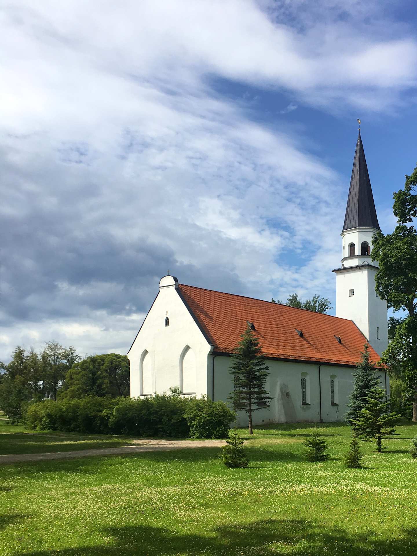 A white church with a tall steeple stands in a lush green field, surrounded by trees and a cloudy blue sky.