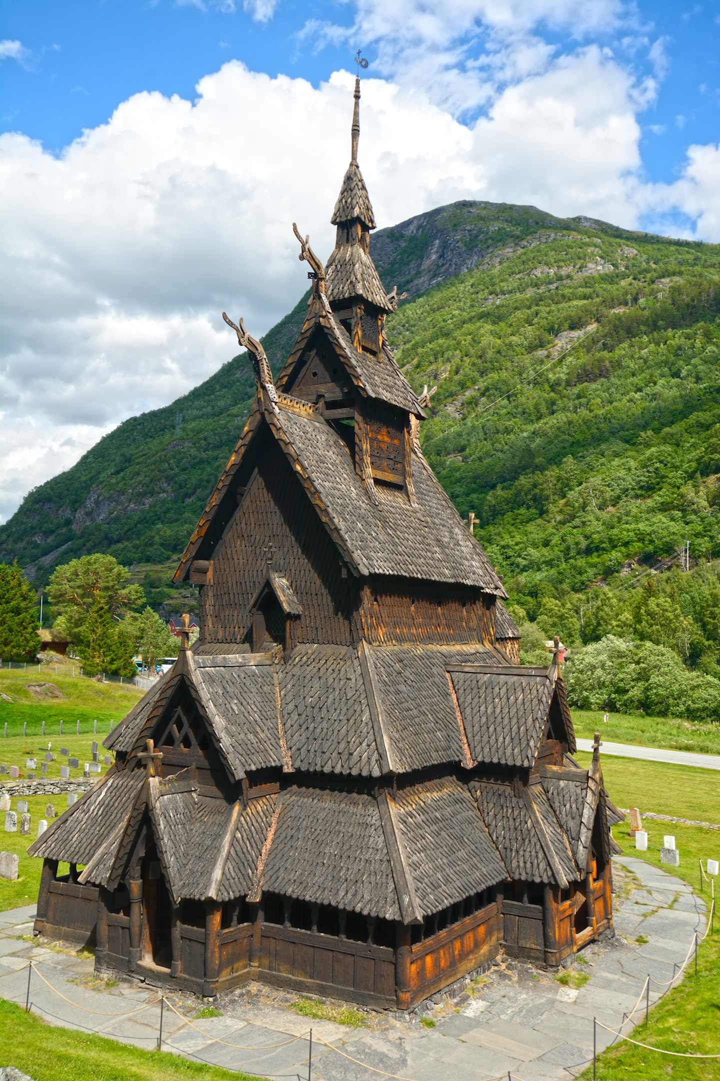 A large, ornate wooden church with a distinctive steeple stands in a lush, green landscape surrounded by mountains.