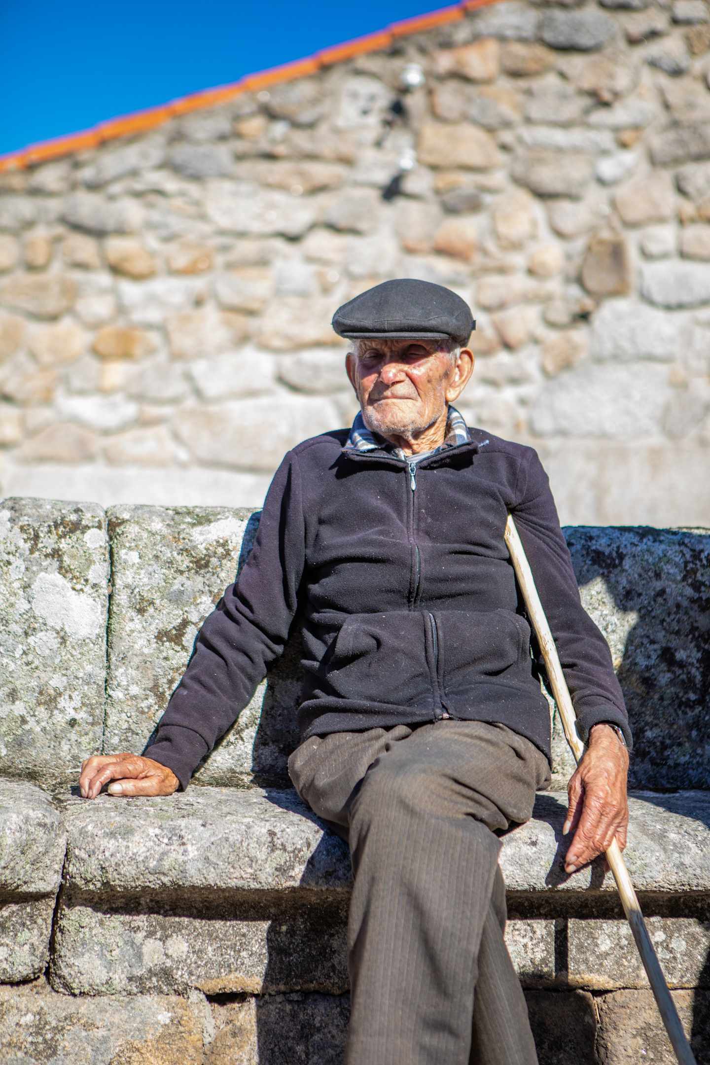 An elderly man wearing a cap and jacket sits on a stone bench in front of a stone wall, with a clear blue sky visible in the background.