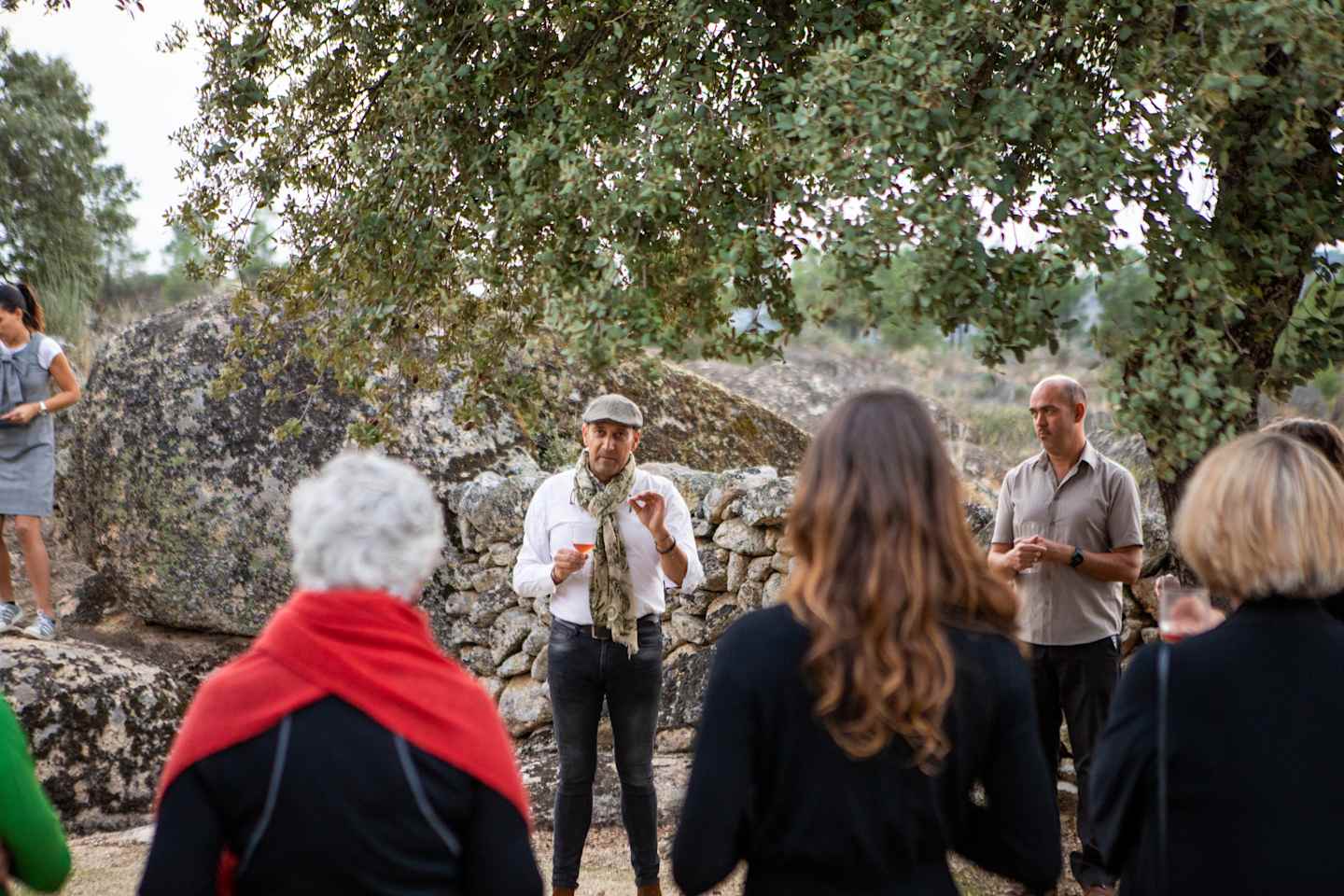 A group of people, some wearing casual clothing and others in more formal attire, are gathered in a wooded area with a stone wall in the background.
