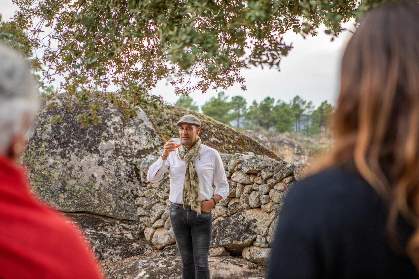 The image shows a person wearing a white shirt and hat standing in front of a stone wall, with trees and foliage in the background.
