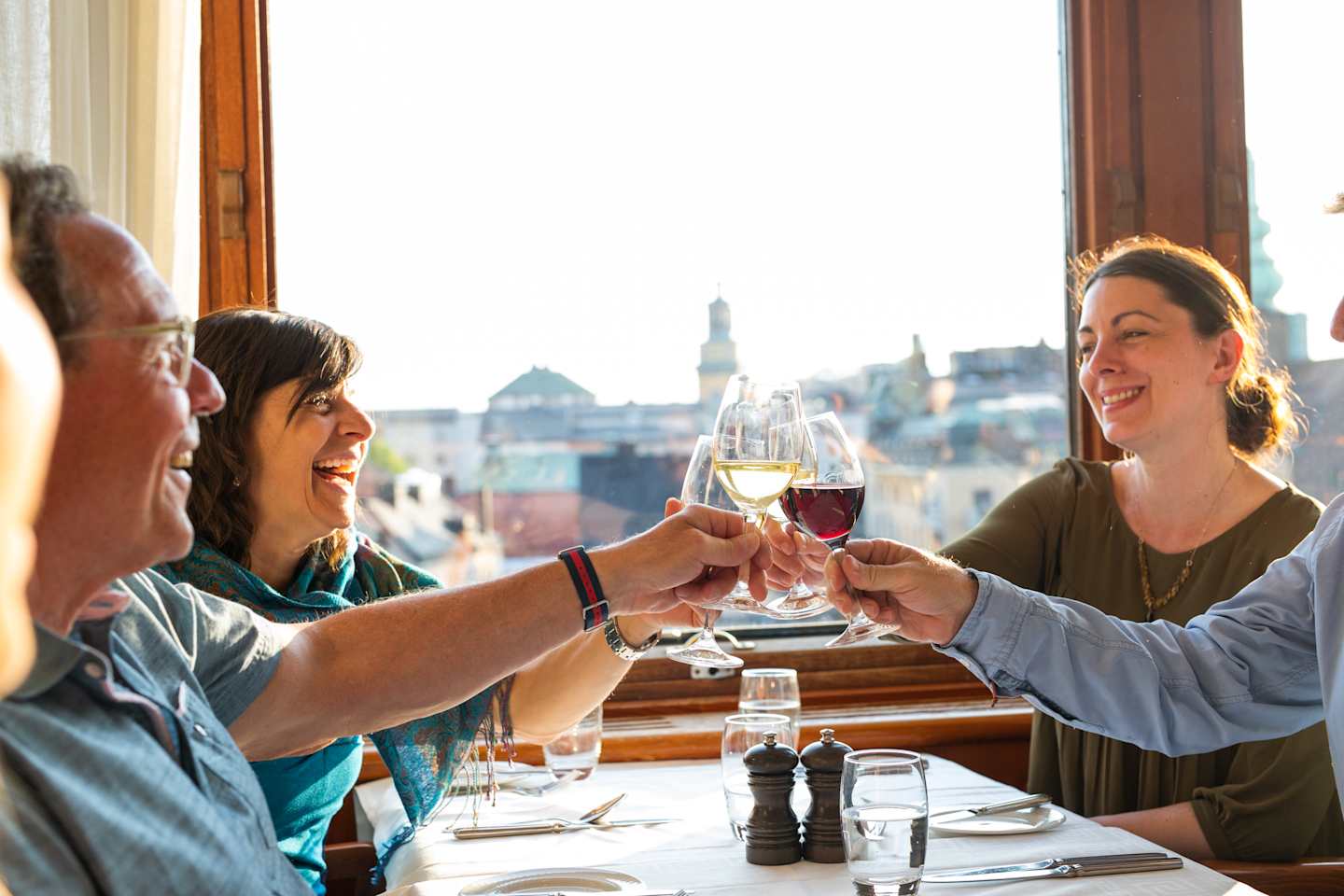 A group of people enjoying drinks and conversation at a table by a window, with a cityscape visible in the background.