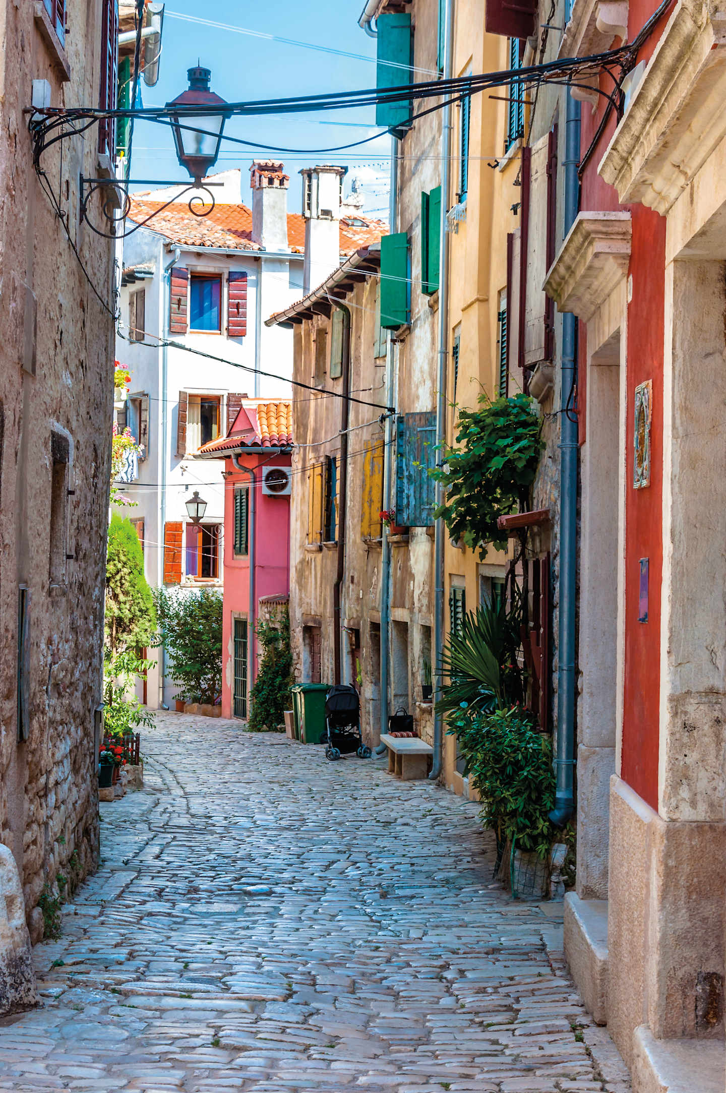 A charming cobblestone alley lined with colorful buildings, potted plants, and hanging laundry, creating a quaint and picturesque scene in what appears to be a historic European town.