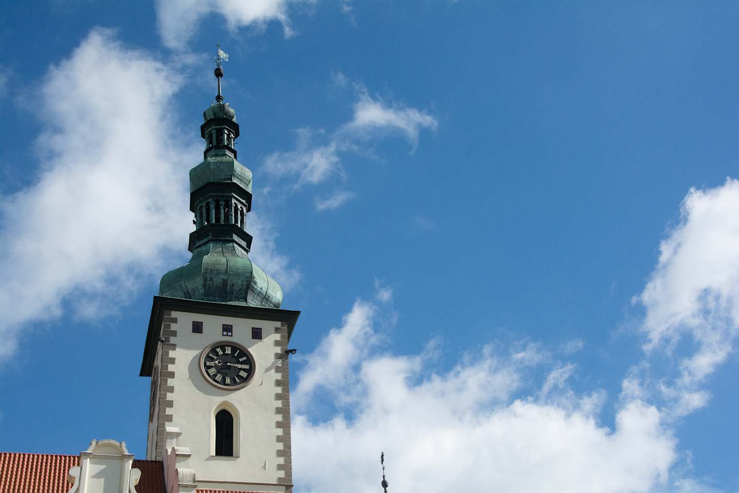 A tall, ornate clock tower with a green copper dome stands against a bright blue sky with fluffy white clouds.