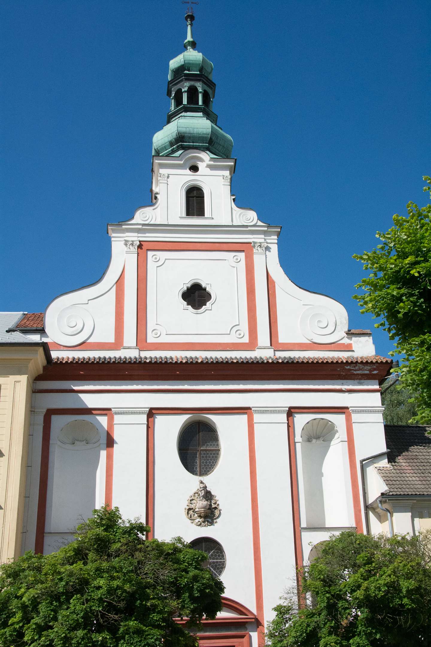 A white and pink ornate church building with a green-domed tower stands against a clear blue sky, surrounded by lush greenery in the foreground.