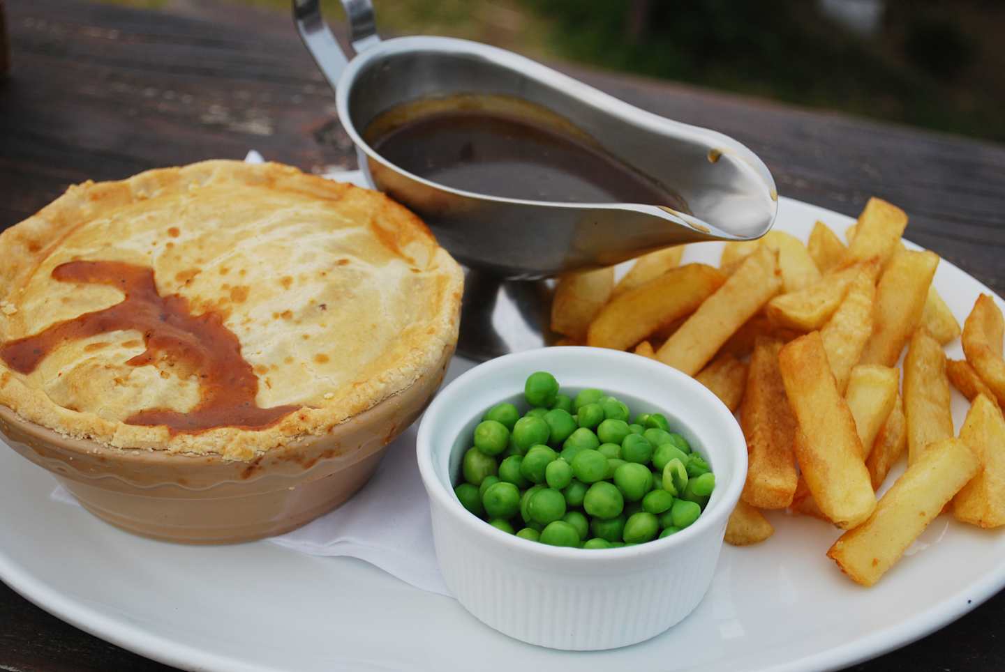 A plate with a savory pie, french fries, and a side of peas, accompanied by a gravy boat, set against a blurred outdoor background.
