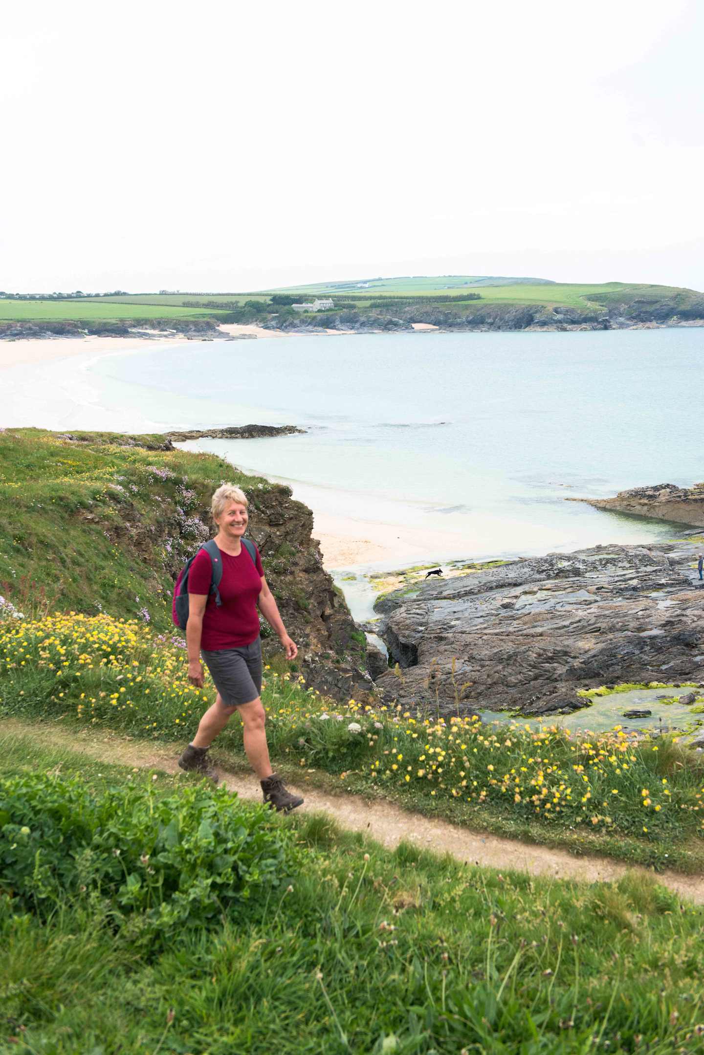 A person in a red shirt and shorts is walking along a grassy path overlooking a scenic coastal landscape with a body of water and distant hills.