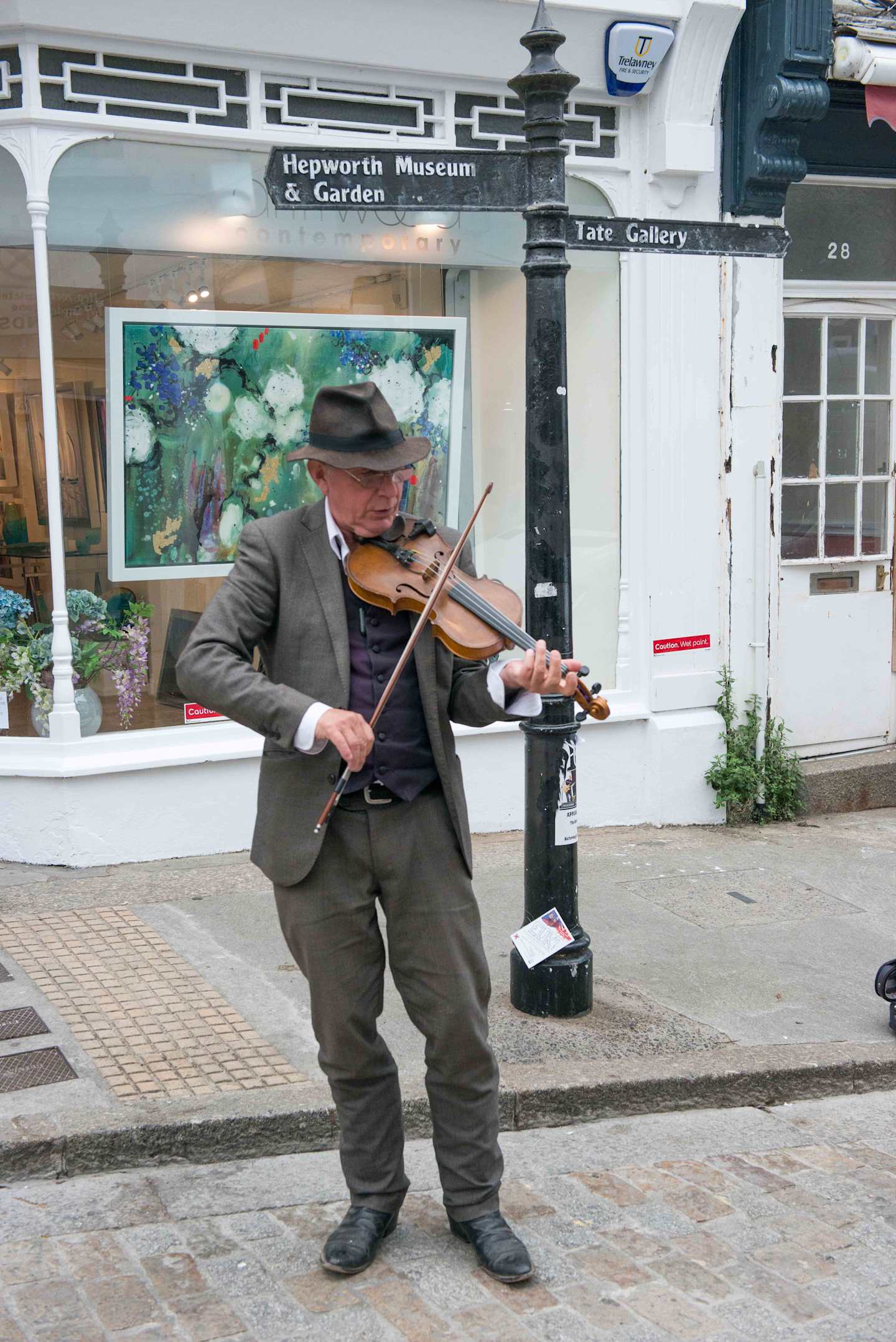 A man in casual attire is playing a violin on the street in front of a museum and art gallery, with a colorful mural visible in the background.
