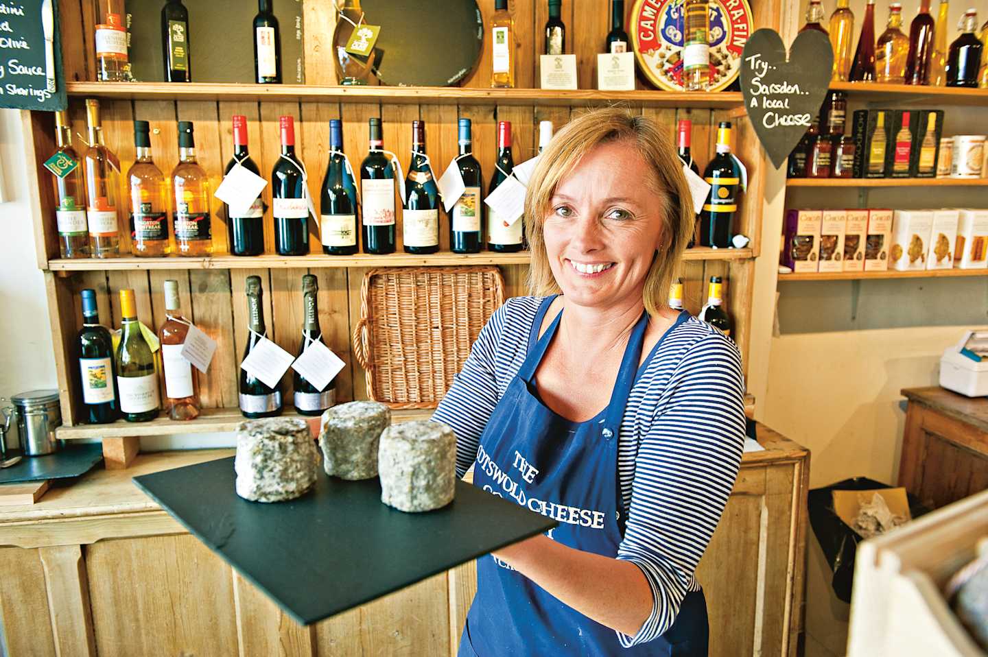 A smiling woman in a blue and white striped shirt stands behind a counter in a wine and cheese shop, surrounded by shelves of wine bottles and other products.