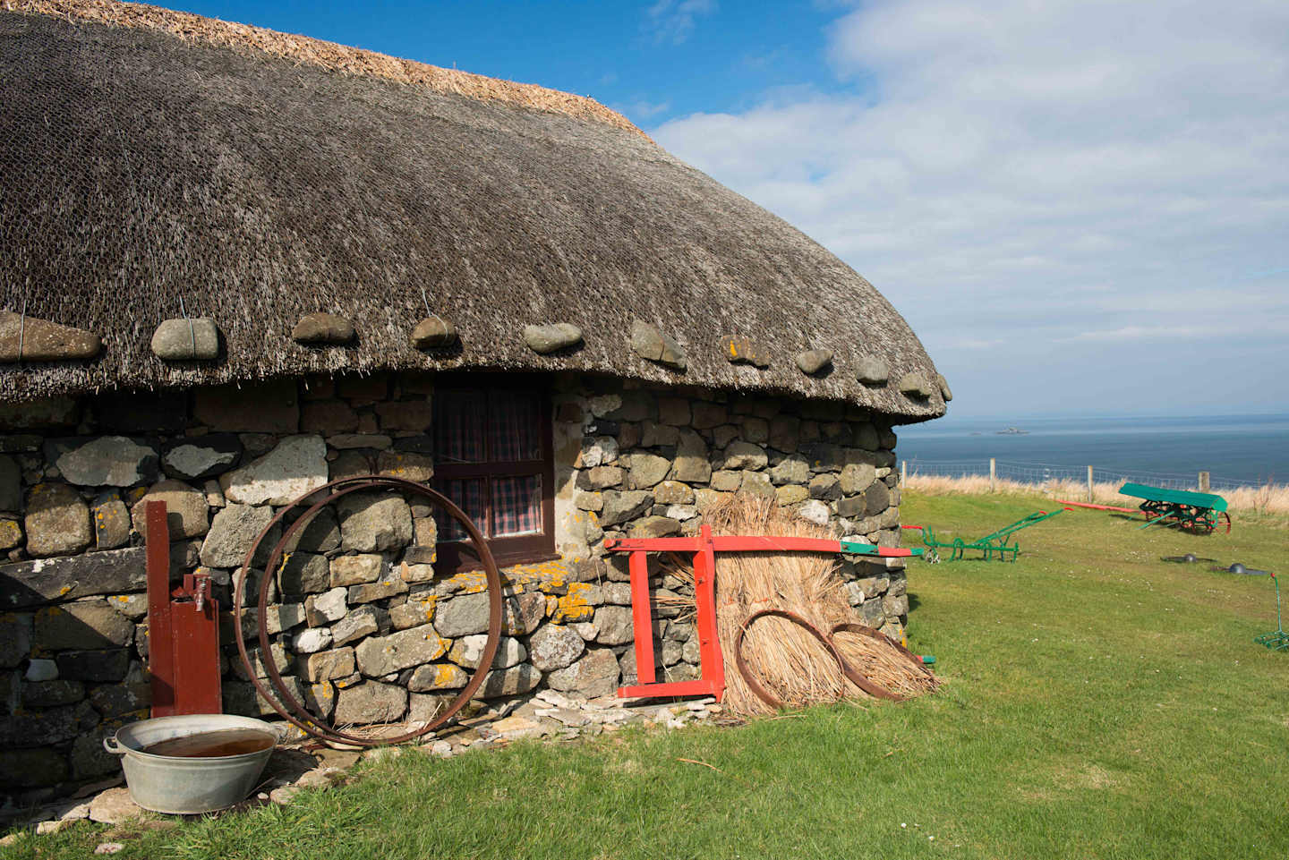 A traditional stone cottage with a thatched roof sits on a grassy hill, overlooking the ocean in the distance.