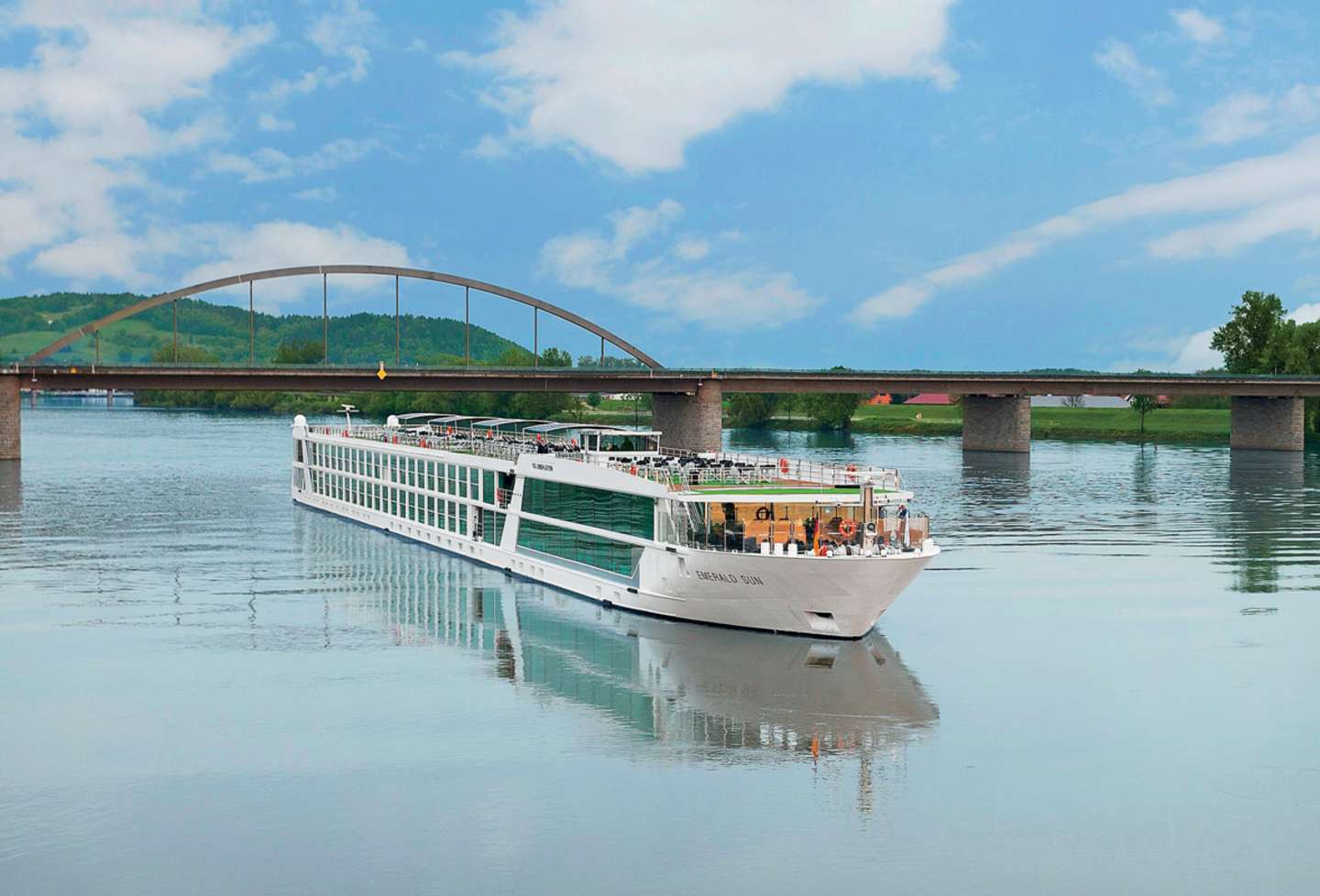 A large passenger boat is sailing on a calm river, with a picturesque arched bridge spanning the waterway in the background against a blue sky with fluffy white clouds.