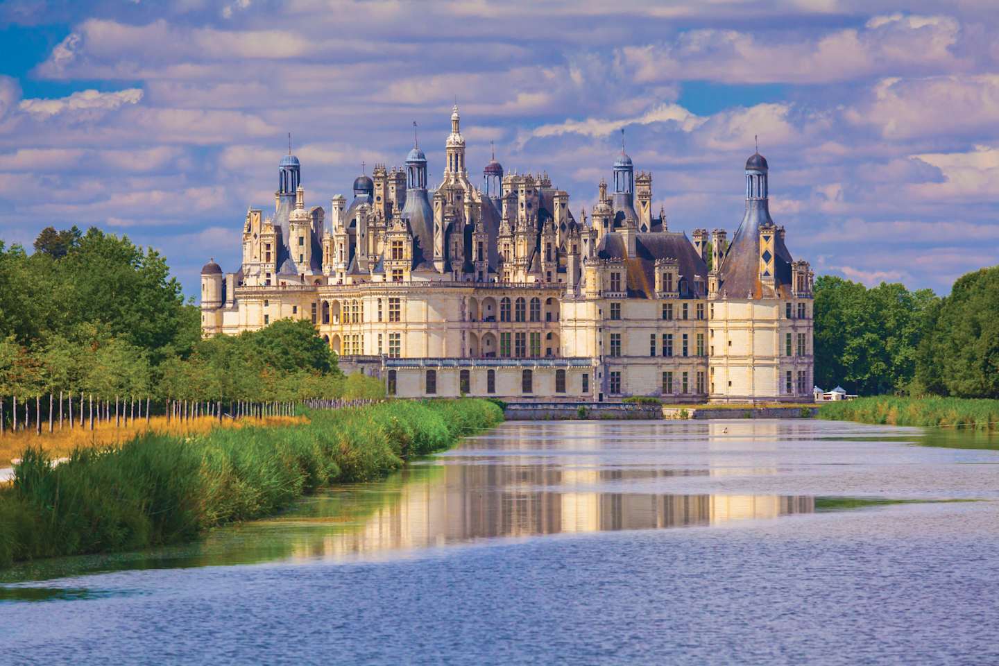 A magnificent Renaissance-style castle with numerous towers and spires stands majestically on the banks of a tranquil body of water, surrounded by lush greenery and a picturesque cloudy sky.