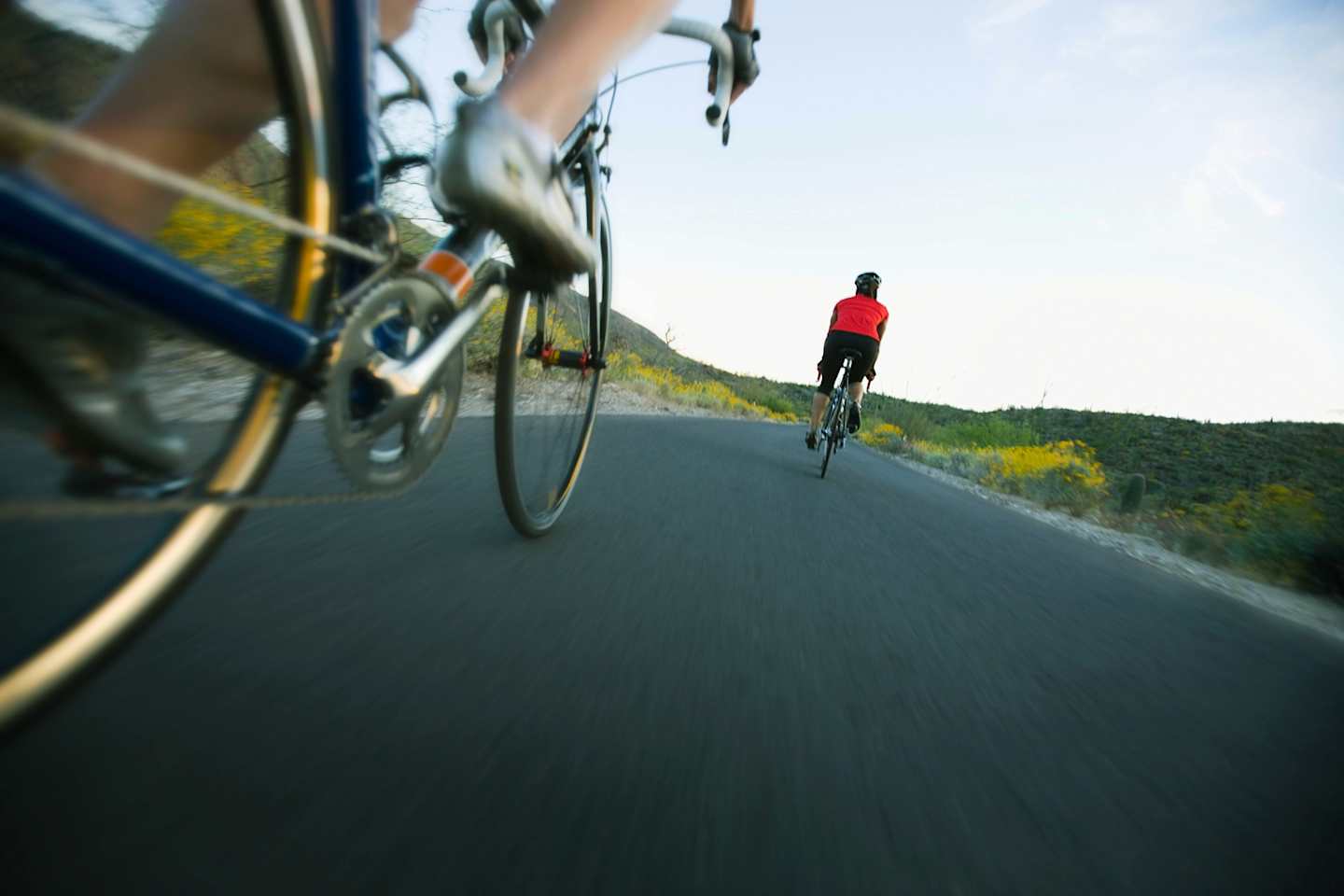 A cyclist rides on a winding road surrounded by lush, hilly terrain in the background.