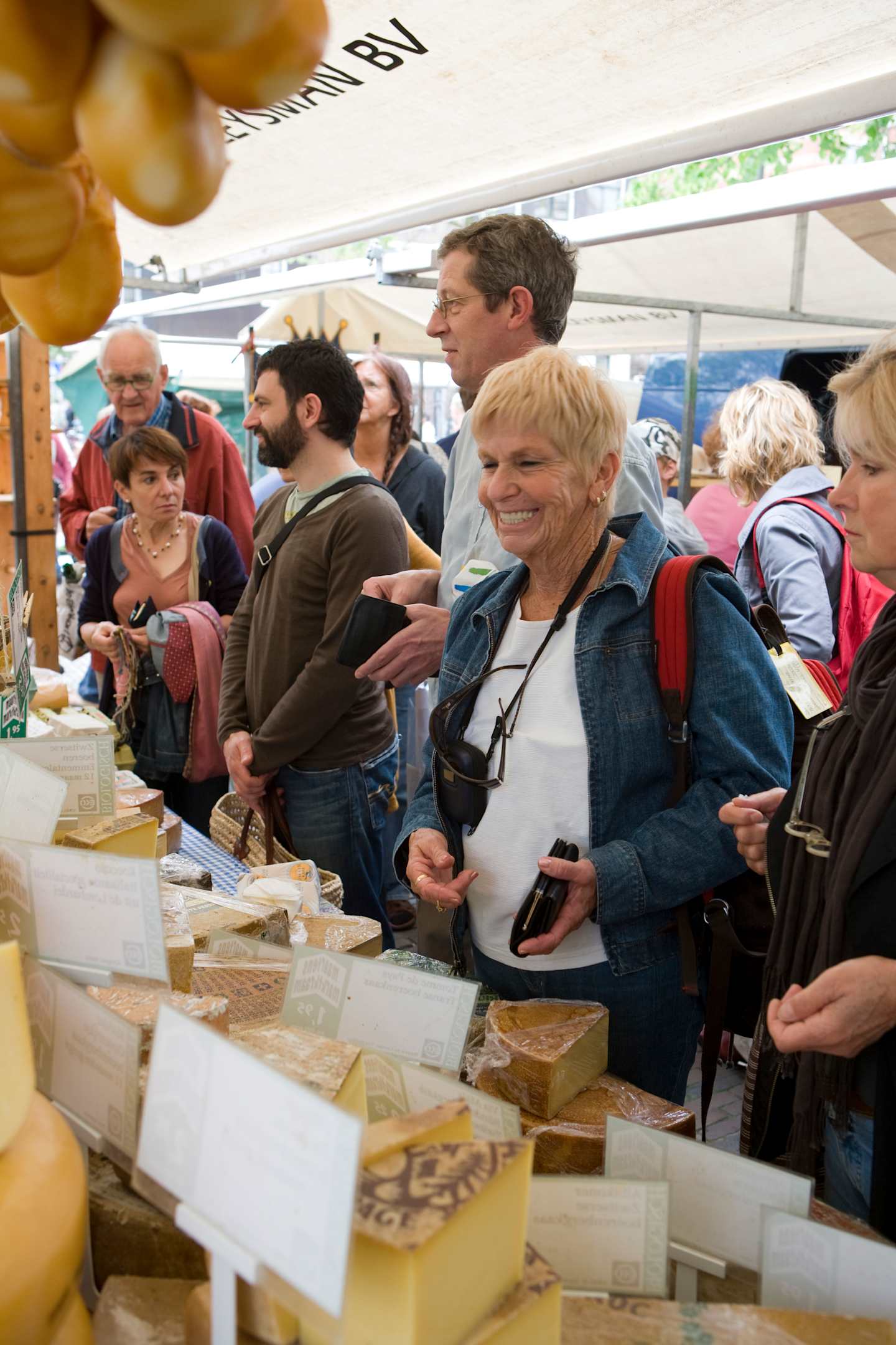 A group of people browsing and purchasing various items from a market stall, with a variety of products displayed on the table in the foreground.