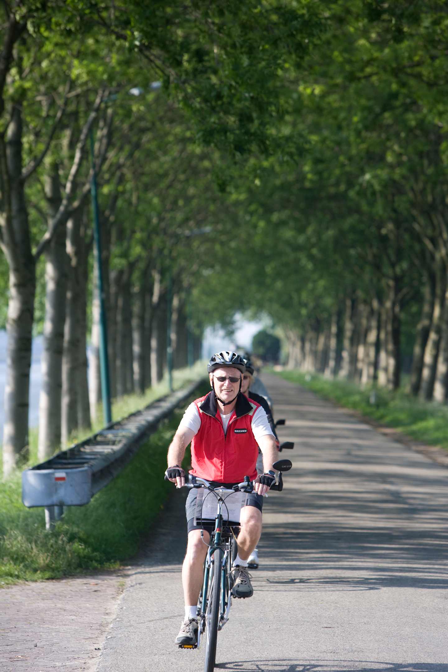 A person in a red jacket and helmet is riding a bicycle down a paved path surrounded by a lush, green forest.