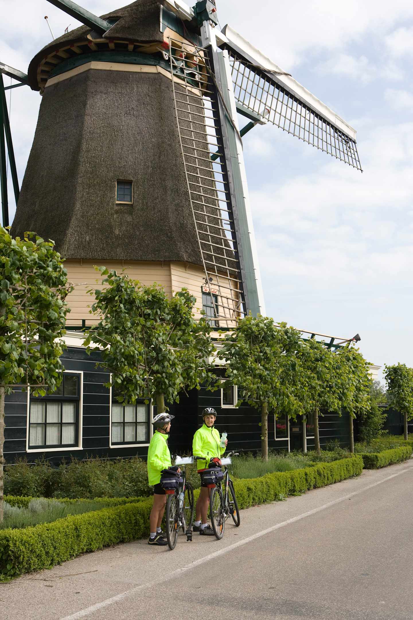 A traditional Dutch windmill stands in the background, surrounded by lush greenery, while two cyclists in bright safety vests ride along the path in the foreground.