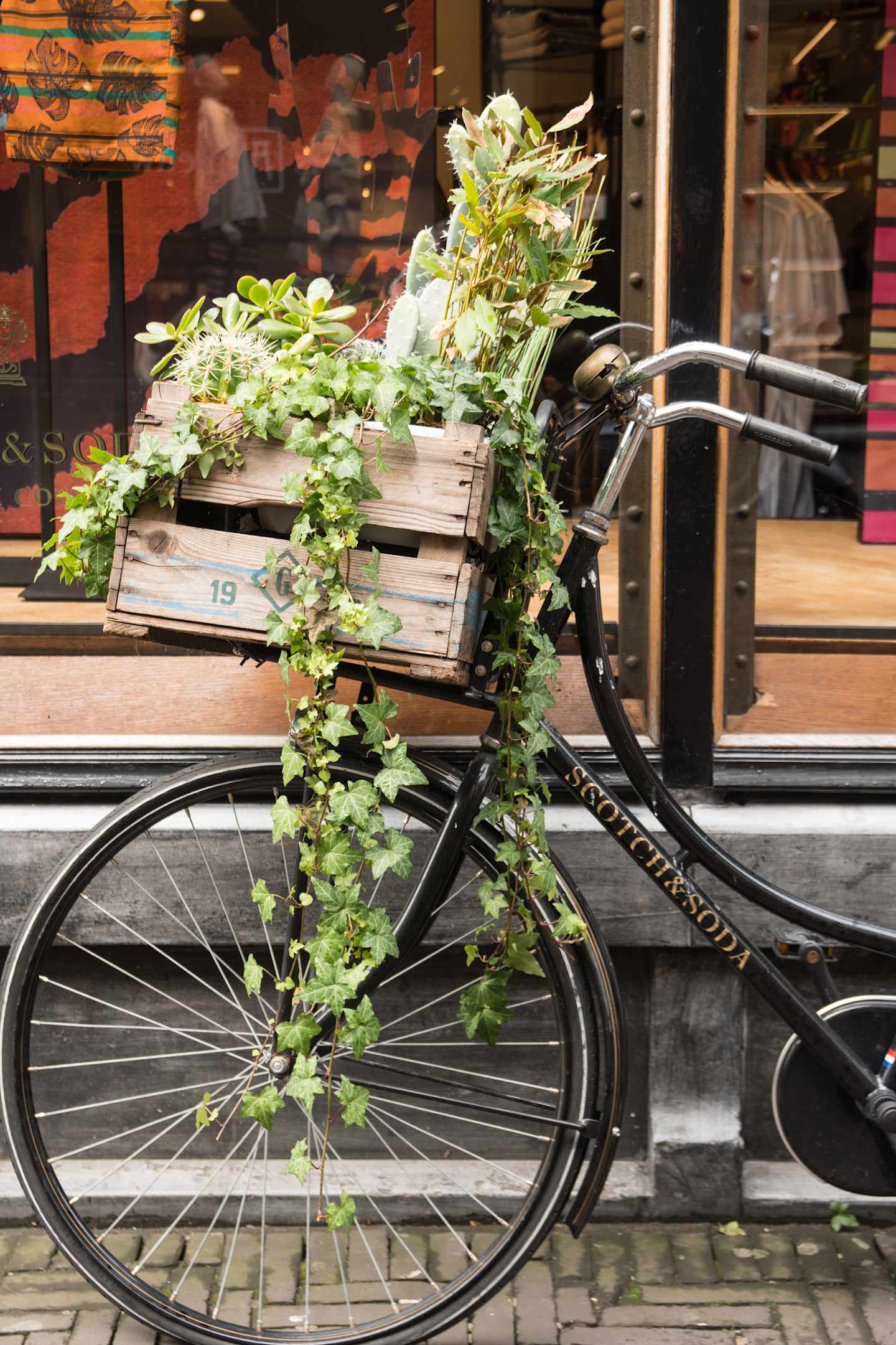 A vintage bicycle with a wooden crate filled with lush, green vines and plants, parked in front of a brick building with a window display.