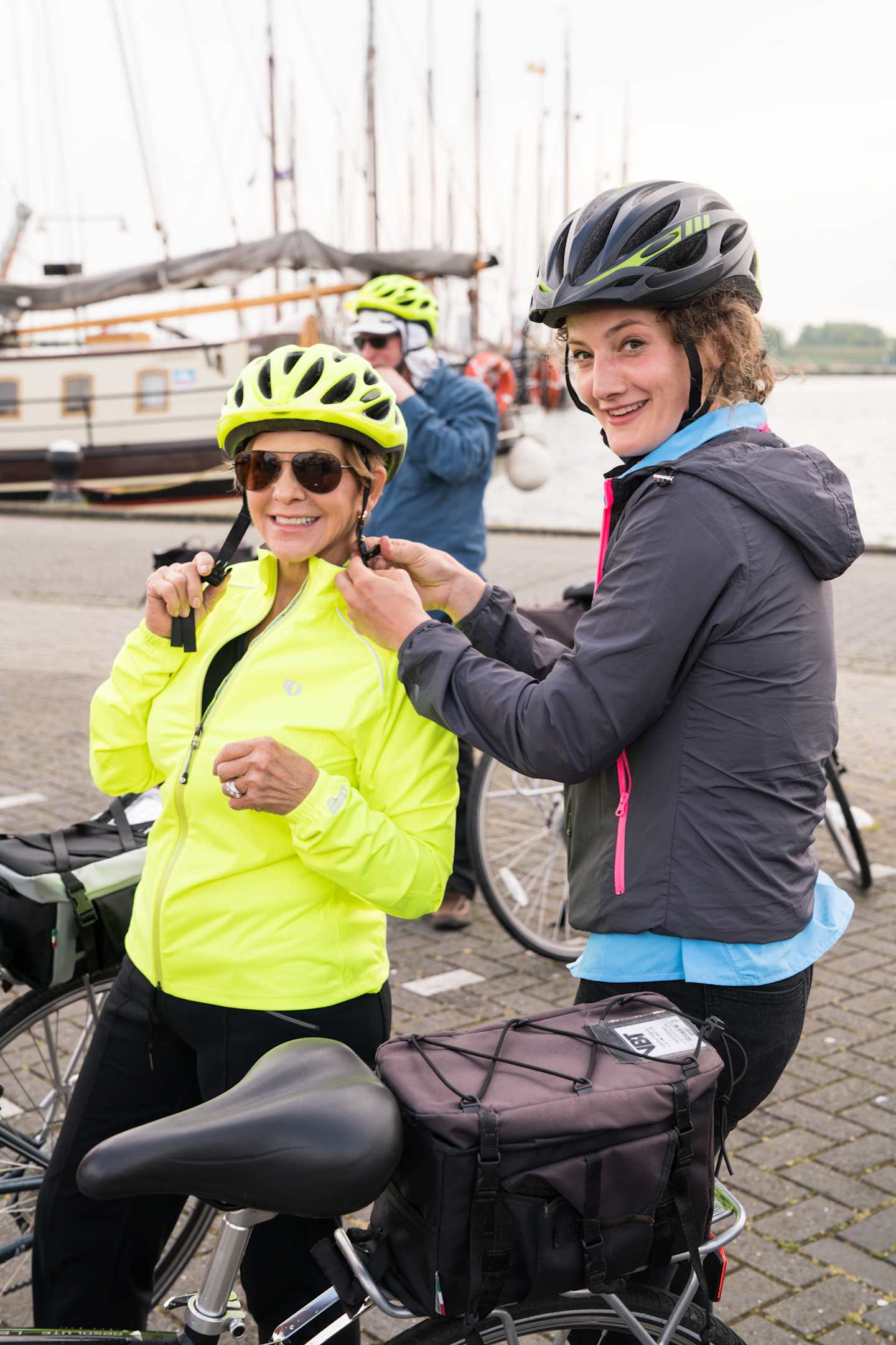 Two women wearing cycling helmets and jackets, smiling and standing in front of a harbor with sailboats in the background.