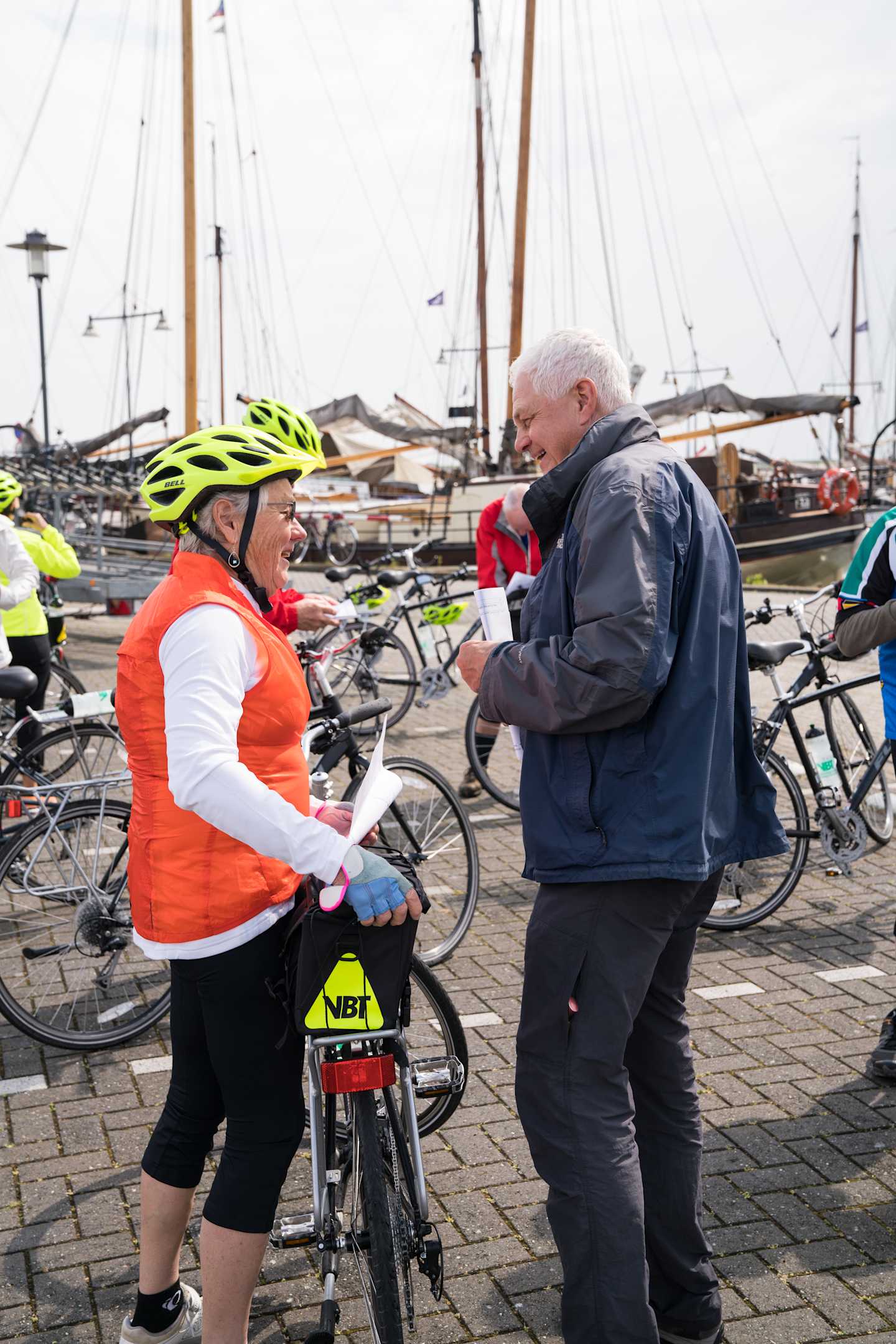The image shows a man and a woman, both wearing cycling gear, engaged in conversation in front of a harbor filled with sailboats.