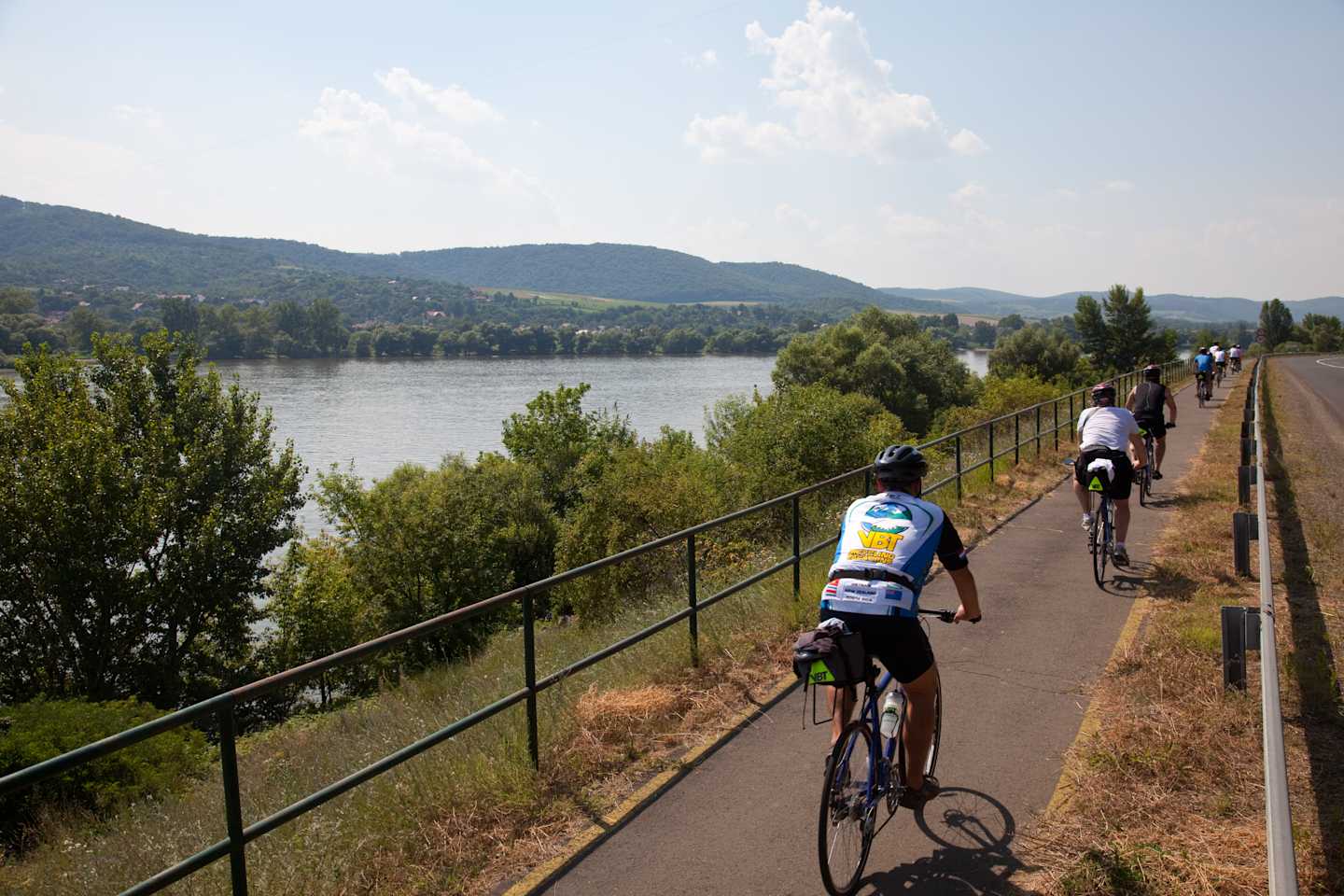A scenic bicycle path runs along the shore of a lake, surrounded by lush greenery and rolling hills in the distance.