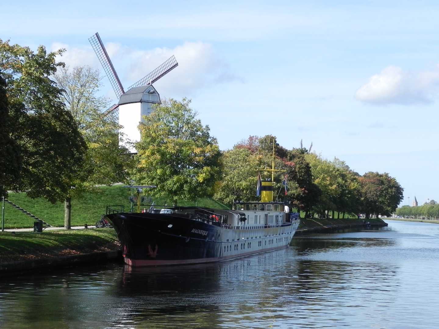 A historic ship sails along a scenic canal surrounded by lush greenery and a traditional windmill in the background.
