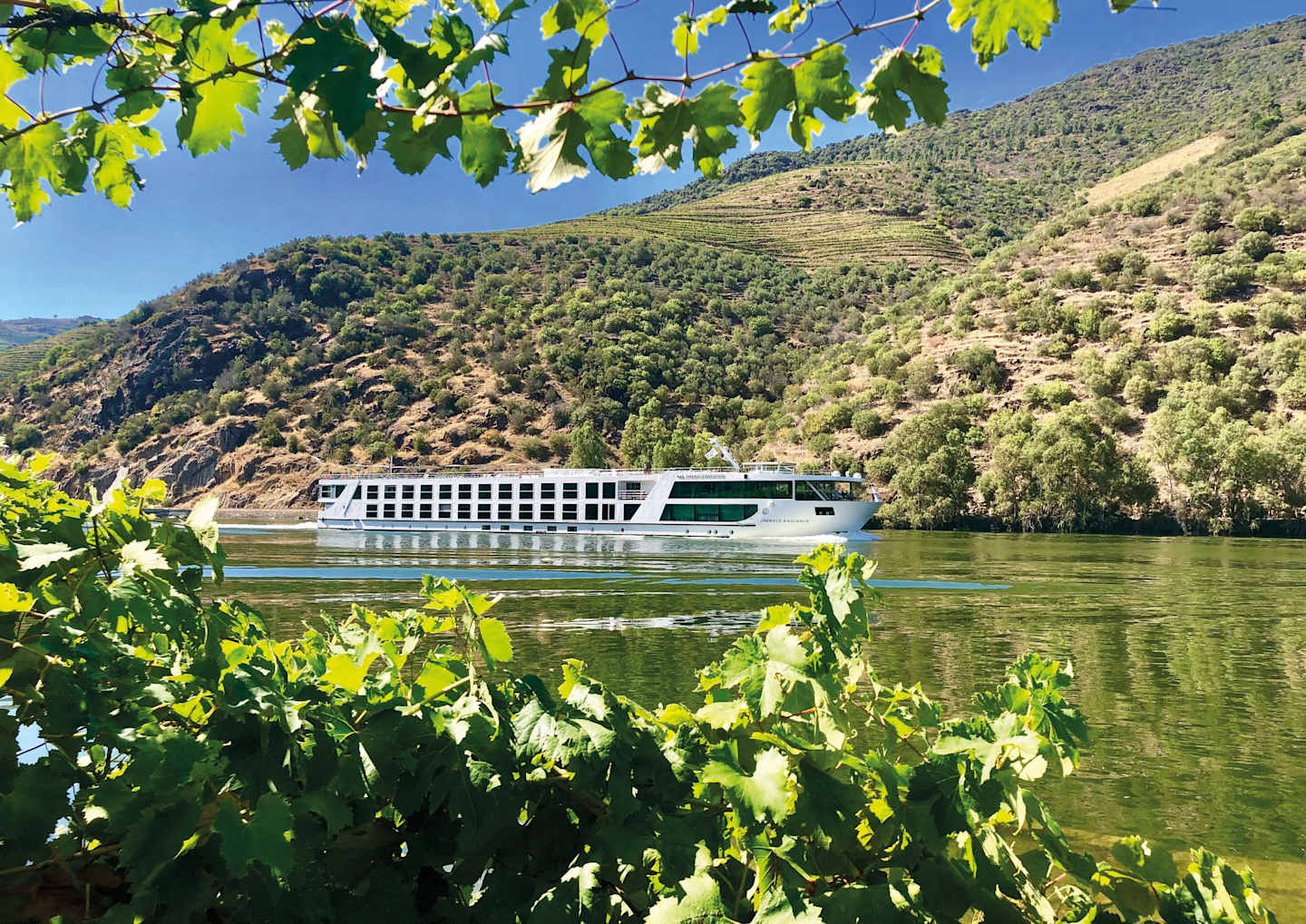 A riverboat cruises along a scenic river surrounded by lush, green foliage and rolling hills in the background.