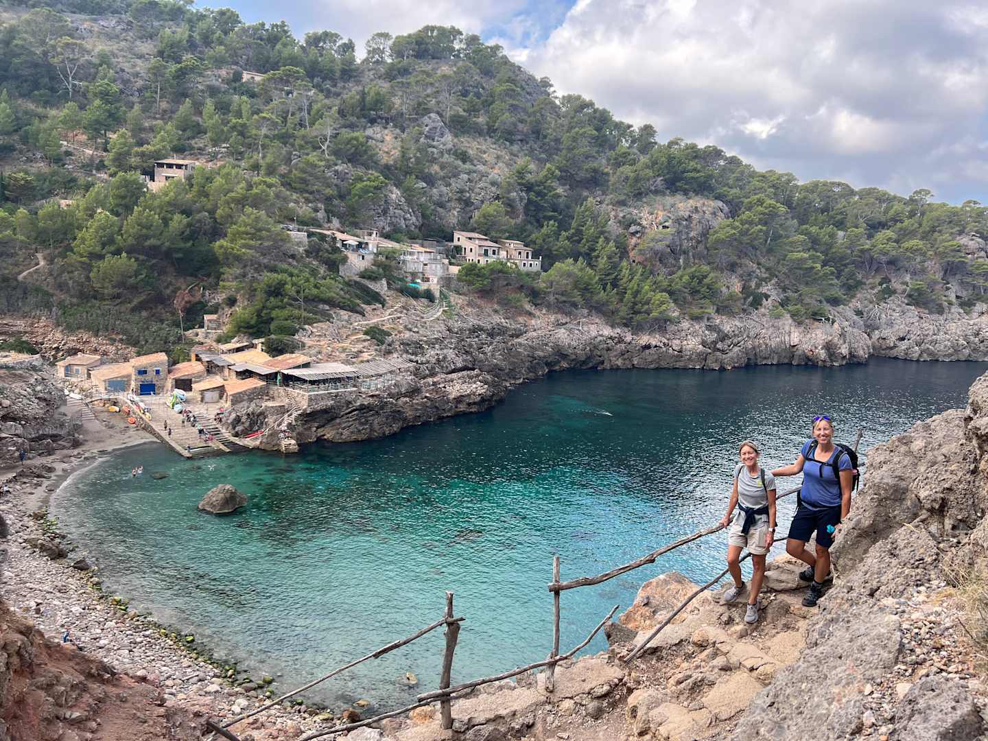 A scenic coastal landscape with a turquoise bay surrounded by rocky cliffs and lush vegetation, with two people walking on a wooden bridge in the foreground.