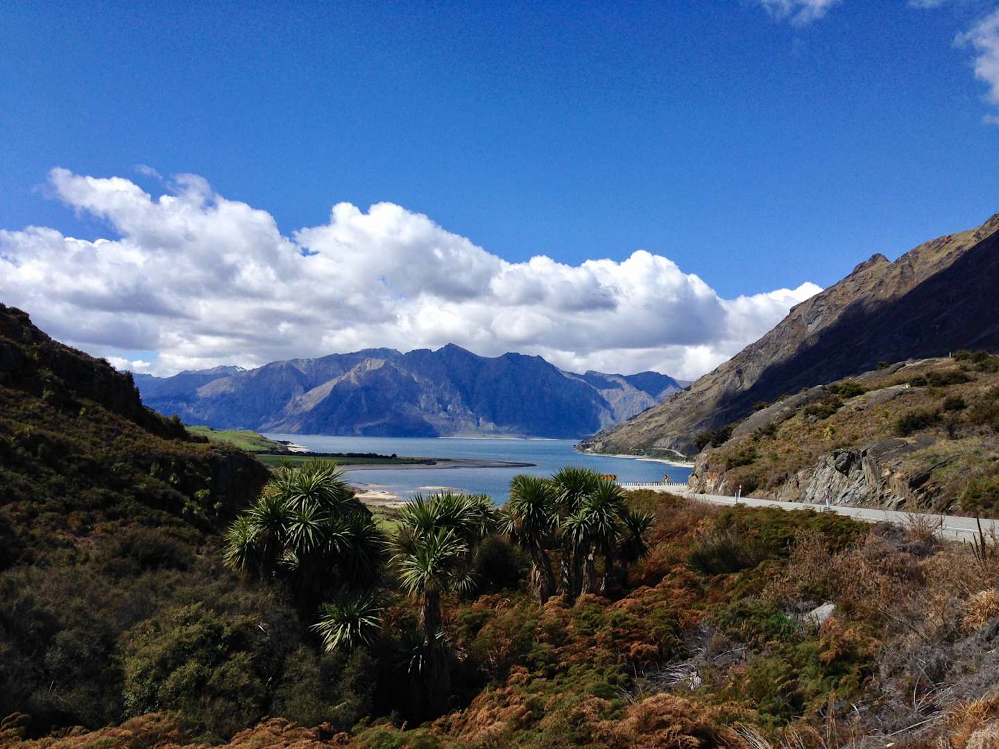 A scenic landscape with a winding road, lush vegetation, and towering mountains in the background, set against a blue sky with fluffy white clouds.